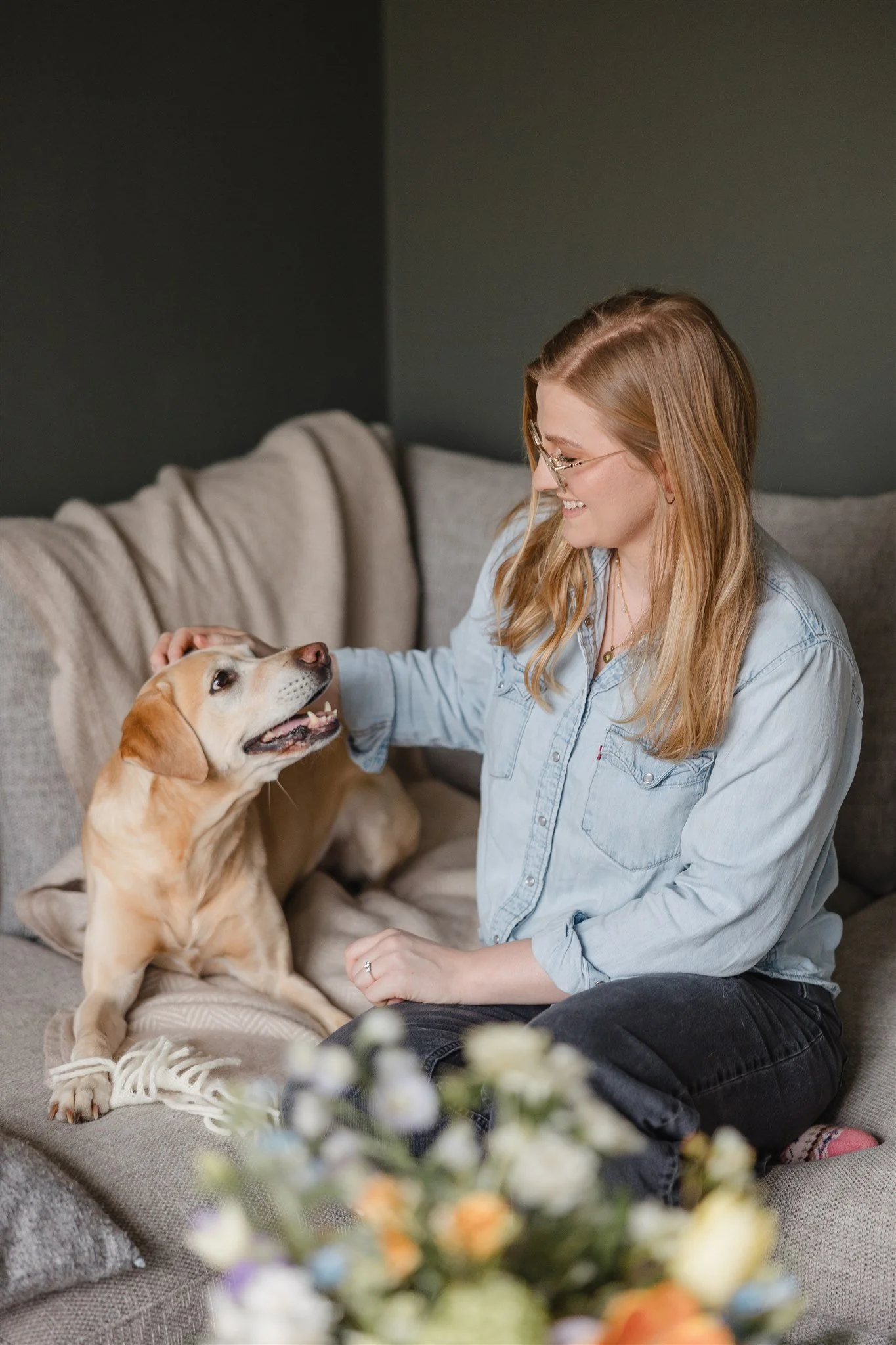 A woman with glasses and long blonde hair smiling and petting a yellow Labrador Retriever dog on a gray couch in a living room.