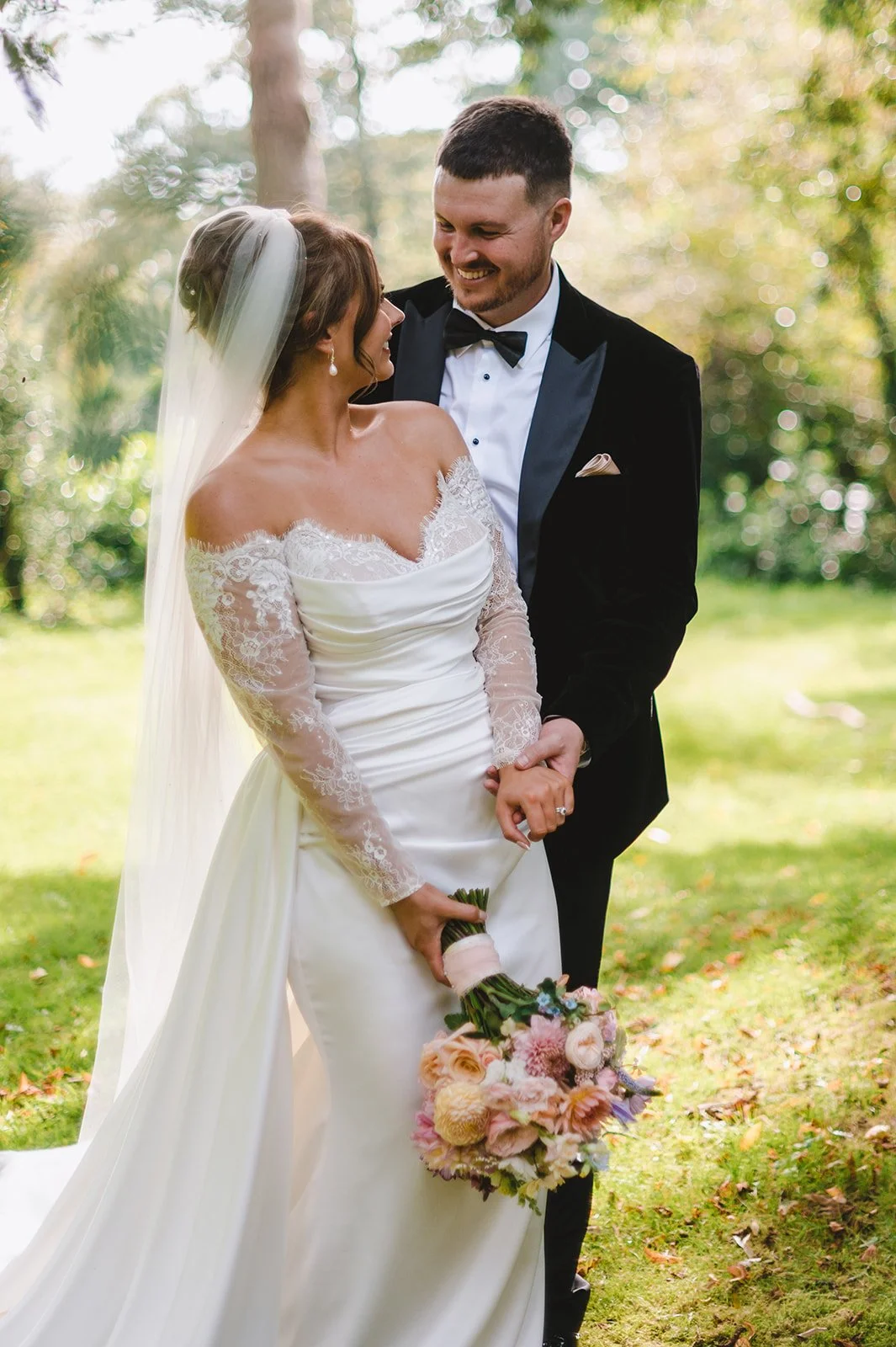 A bride and groom standing outdoors on their wedding day, smiling and holding hands, with the bride holding a bouquet of flowers.