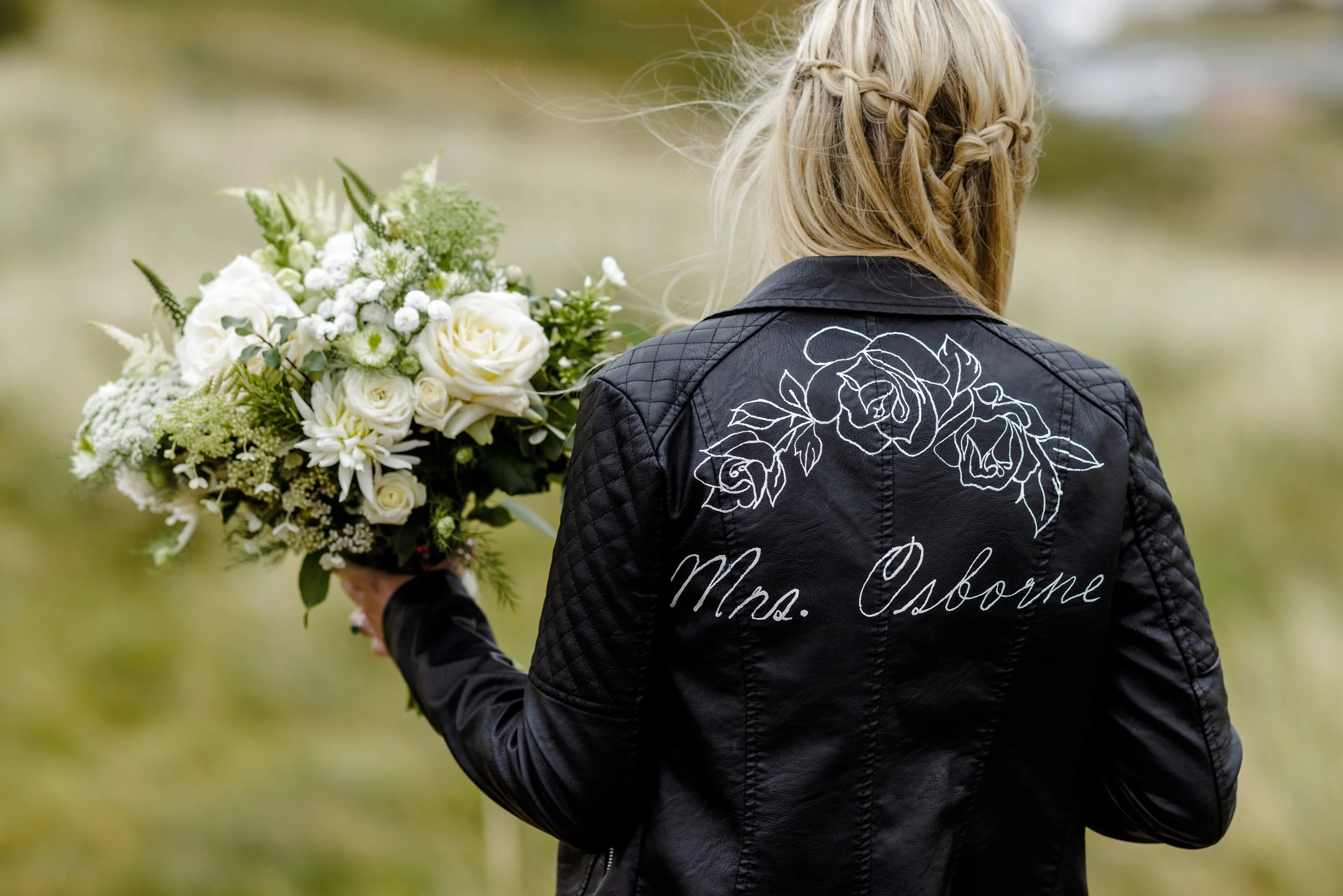Woman with blonde hair in braids wearing a black leather jacket with white floral embroidery and 'Mrs. Osborne' written on the back, holding a large bouquet of white and green flowers outdoors.