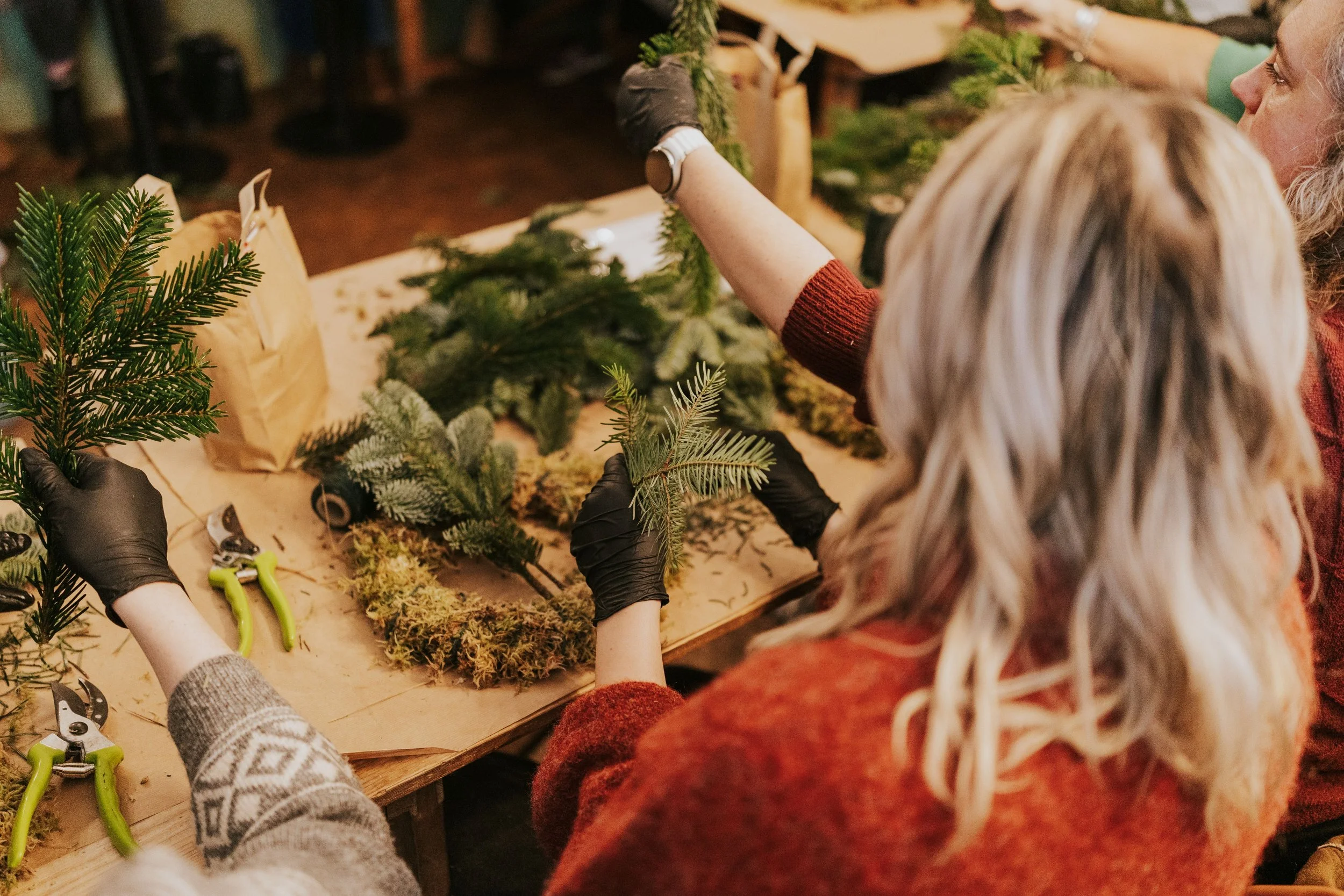 People working on a Christmas wreath craft, arranging green pine branches and moss on a table, with tools and paper bags nearby.
