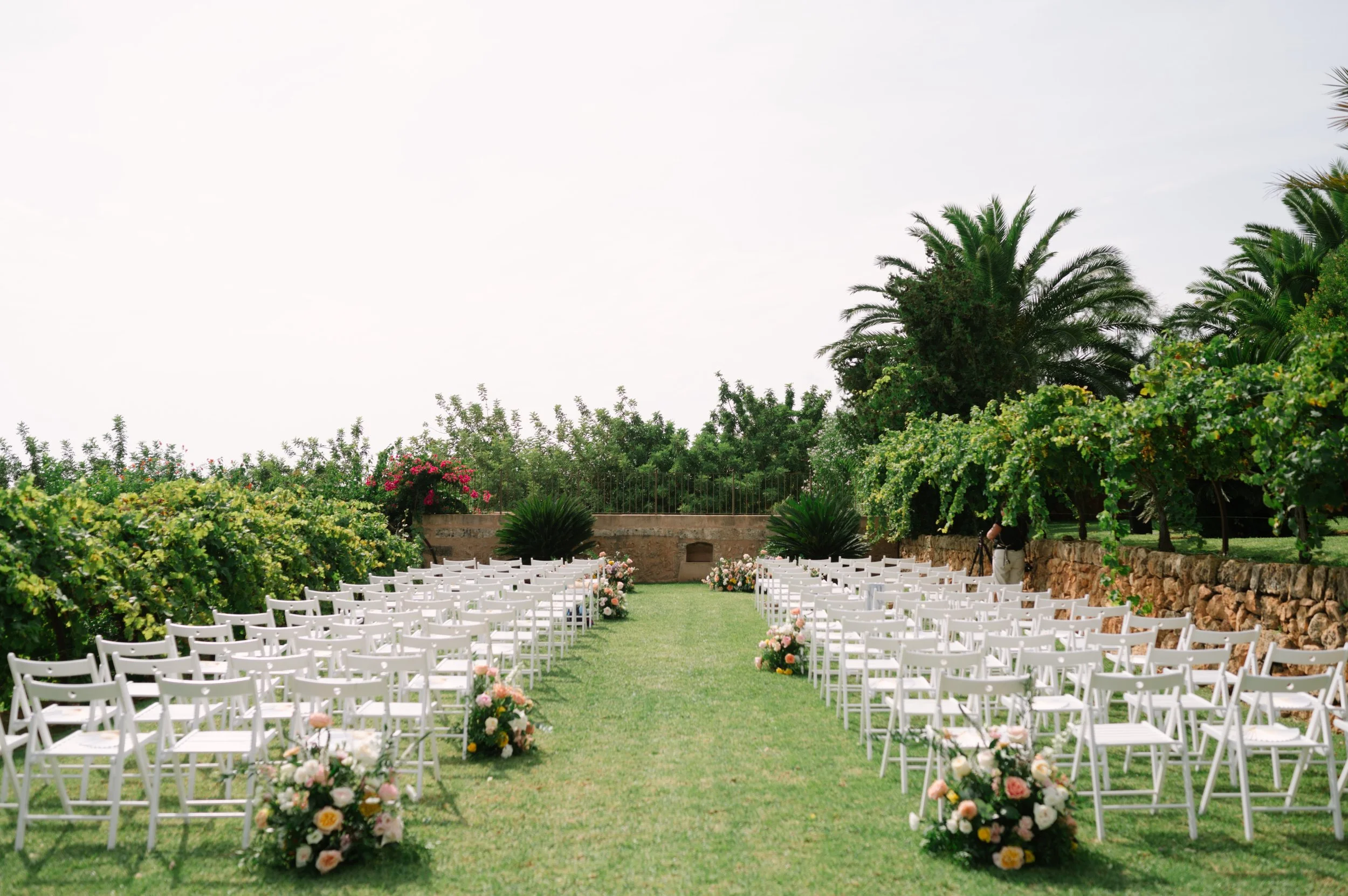 Wedding ceremony setup outdoors with rows of white chairs on a grassy aisle, decorated with pink and white flower arrangements, in a garden with lush green trees and plants.