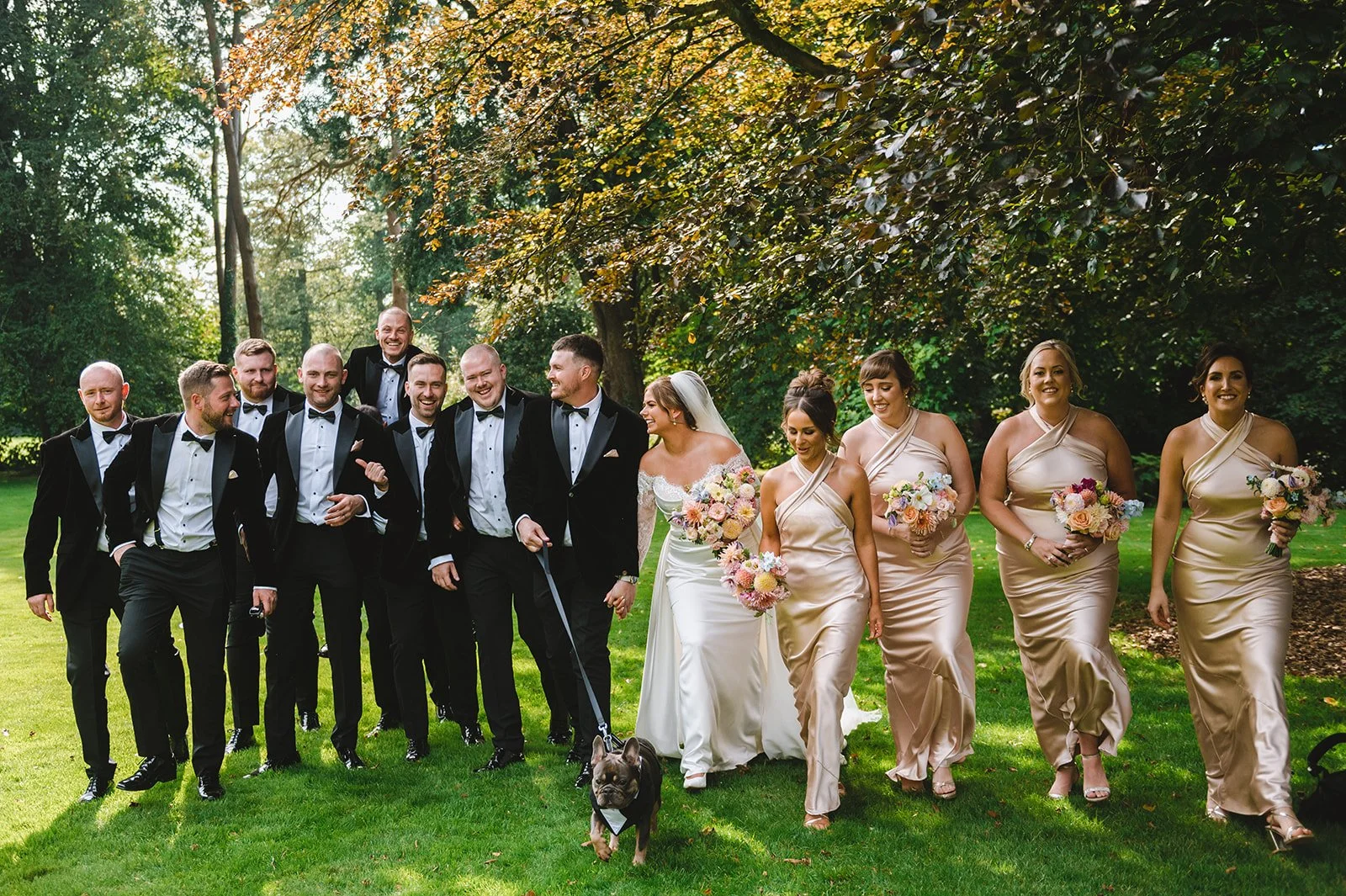 Group of people in wedding attire walking outdoors on a grassy area with trees in the background, including a bride with a veil and bouquet, bridesmaids with bouquets, and groomsmen in tuxedos, along with a small dog on a leash.