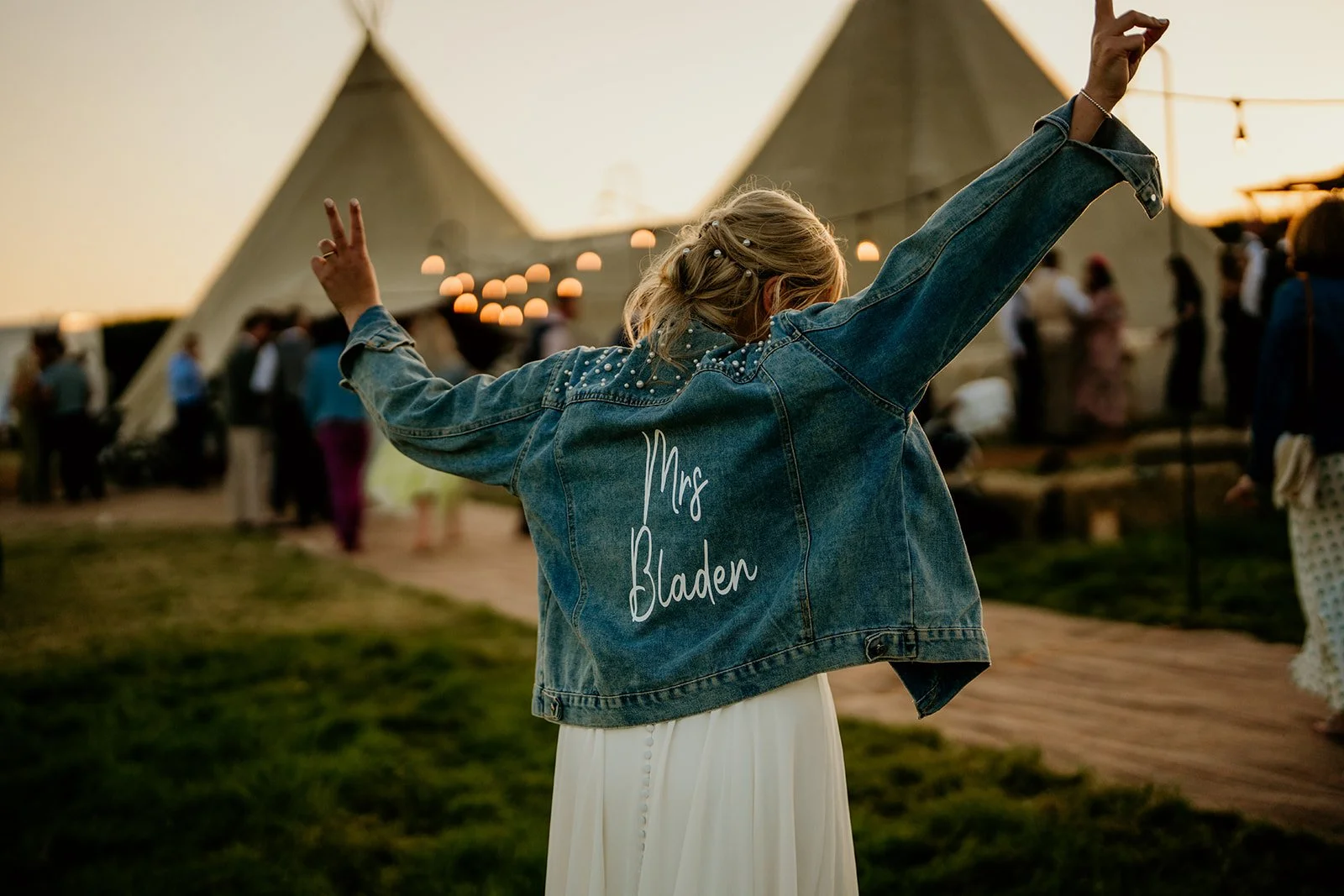 A woman wearing a denim jacket with 'Mrs. Bladen' embroidered on the back is dancing at an outdoor event during sunset, with tents and people in the background.