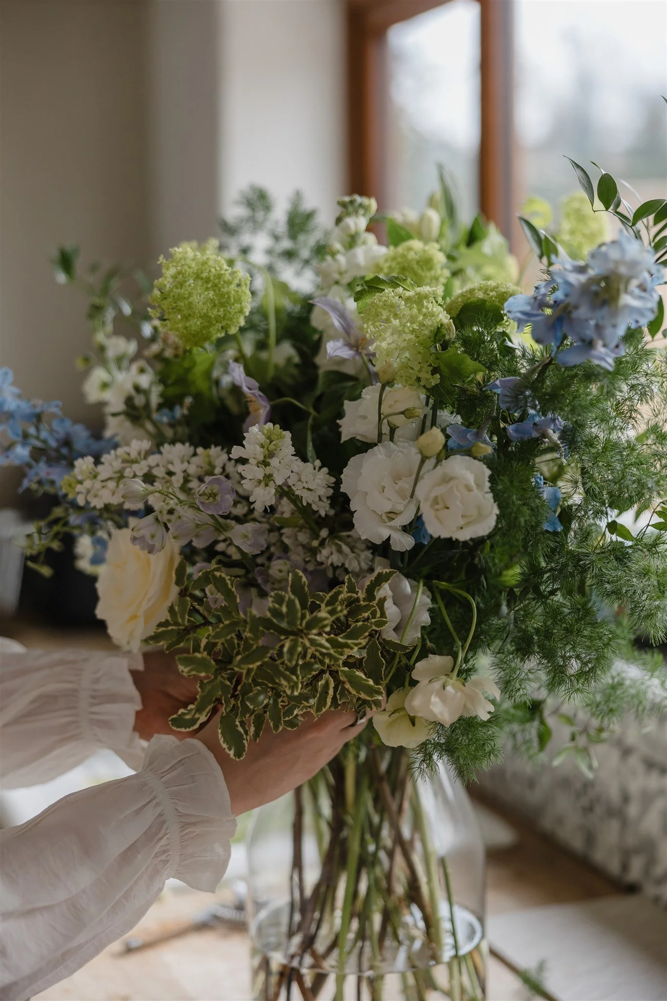 a colourful bouquet of white, blue and green flowers and foliages including roses and delphiniums being arranged in a glass vase.