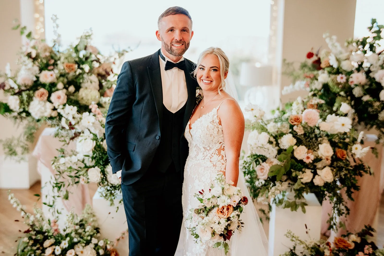 Bride and groom smiling directly at the camera surrounded by an abundance of flowers in whites, pinks and champagne tones. Groom is wearing a black tux with one hand in his pocket, bride is wearing a white lace dress and is holding a bouquet of flowe