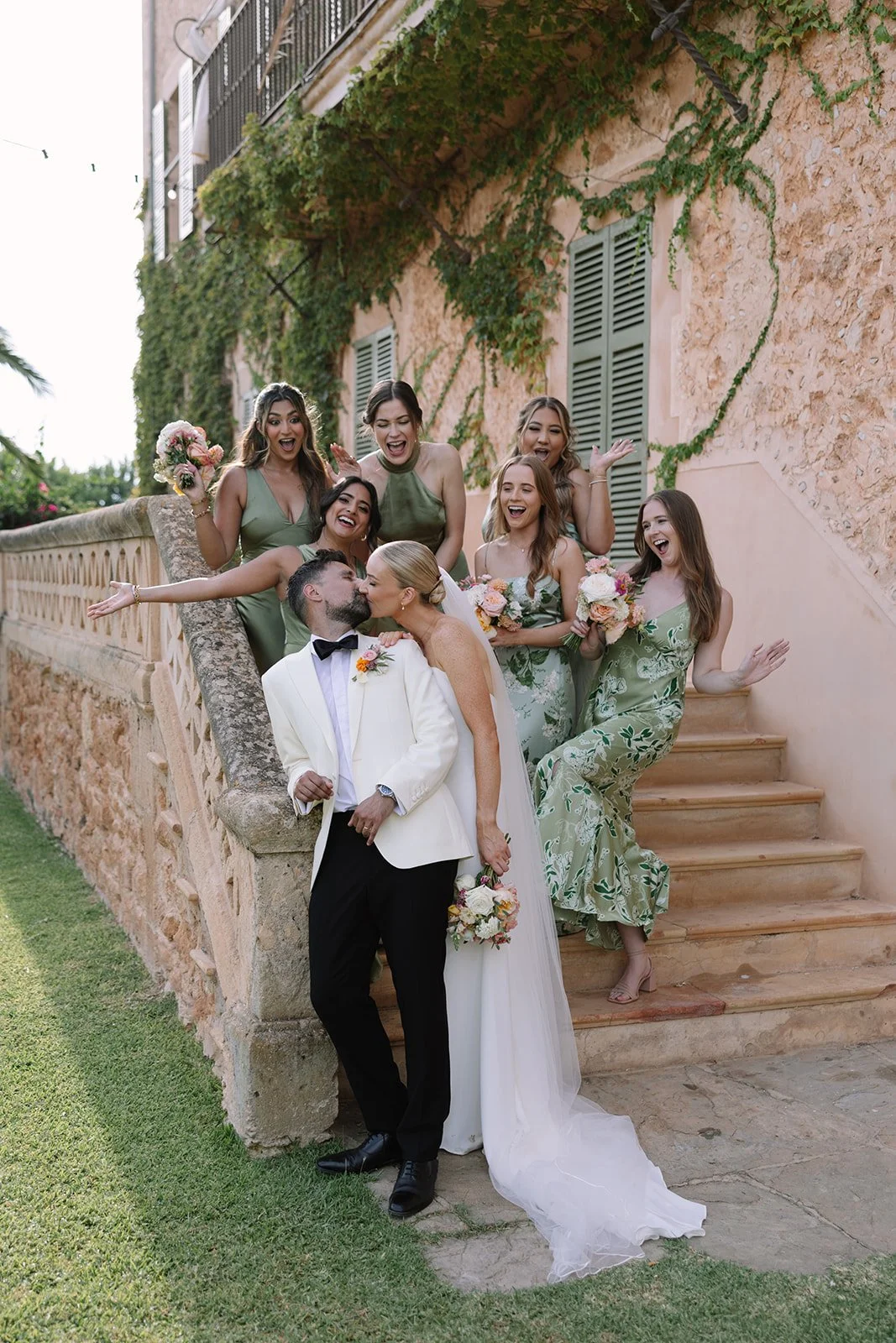 A wedding celebration with a bride and groom kissing in front of friends, surrounded by a stone staircase and a building with ivy-covered walls.
