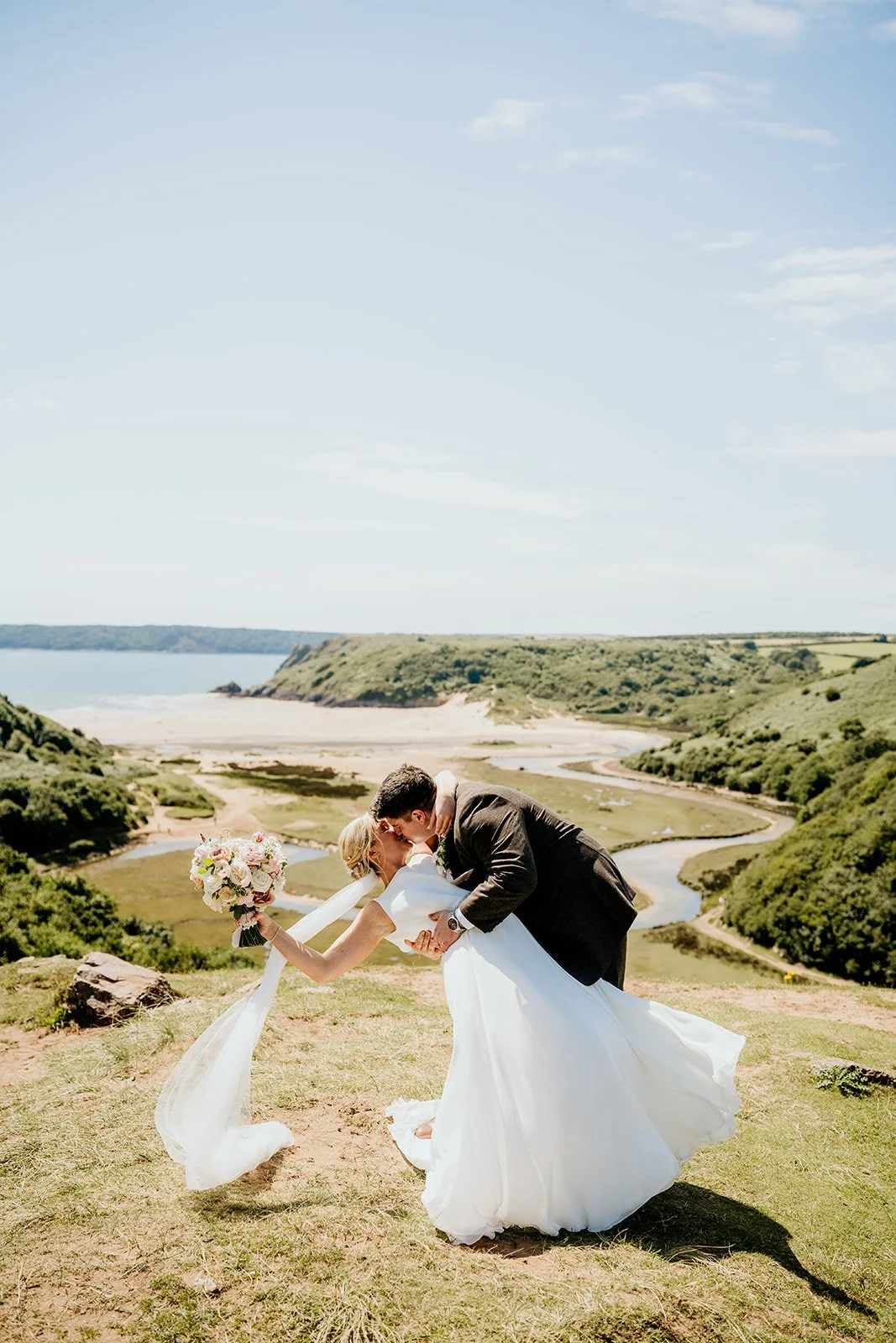 bride and groom kissing with a gorgeous background of three cliff bay, hills, tress and  the beach in the background. Bride is holding  large bouquet of white and pink flowers wearing a white floaty dress. her veil blowing in the wind. 