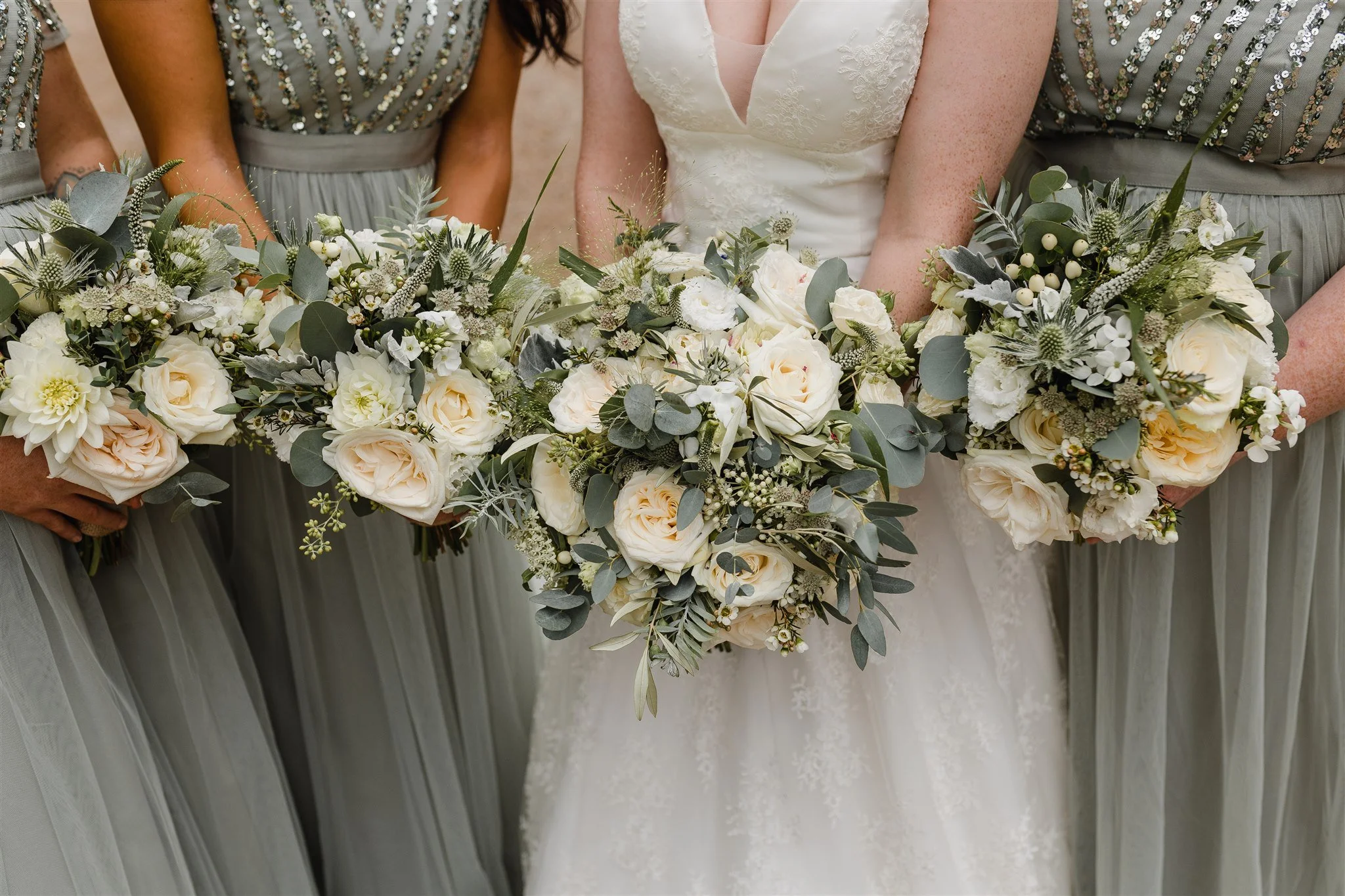 Close-up of a bride and her bridesmaids holding bouquets of white and cream-colored roses, along with greenery with silver leaves, during a wedding.