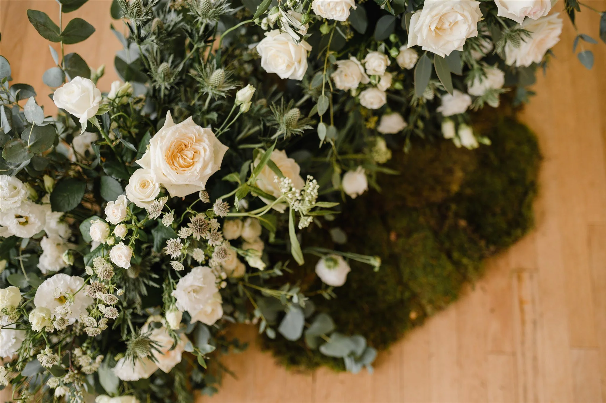 Close-up view of a floral arrangement with white roses, white lisianthus, greenery, and moss on a wooden floor.