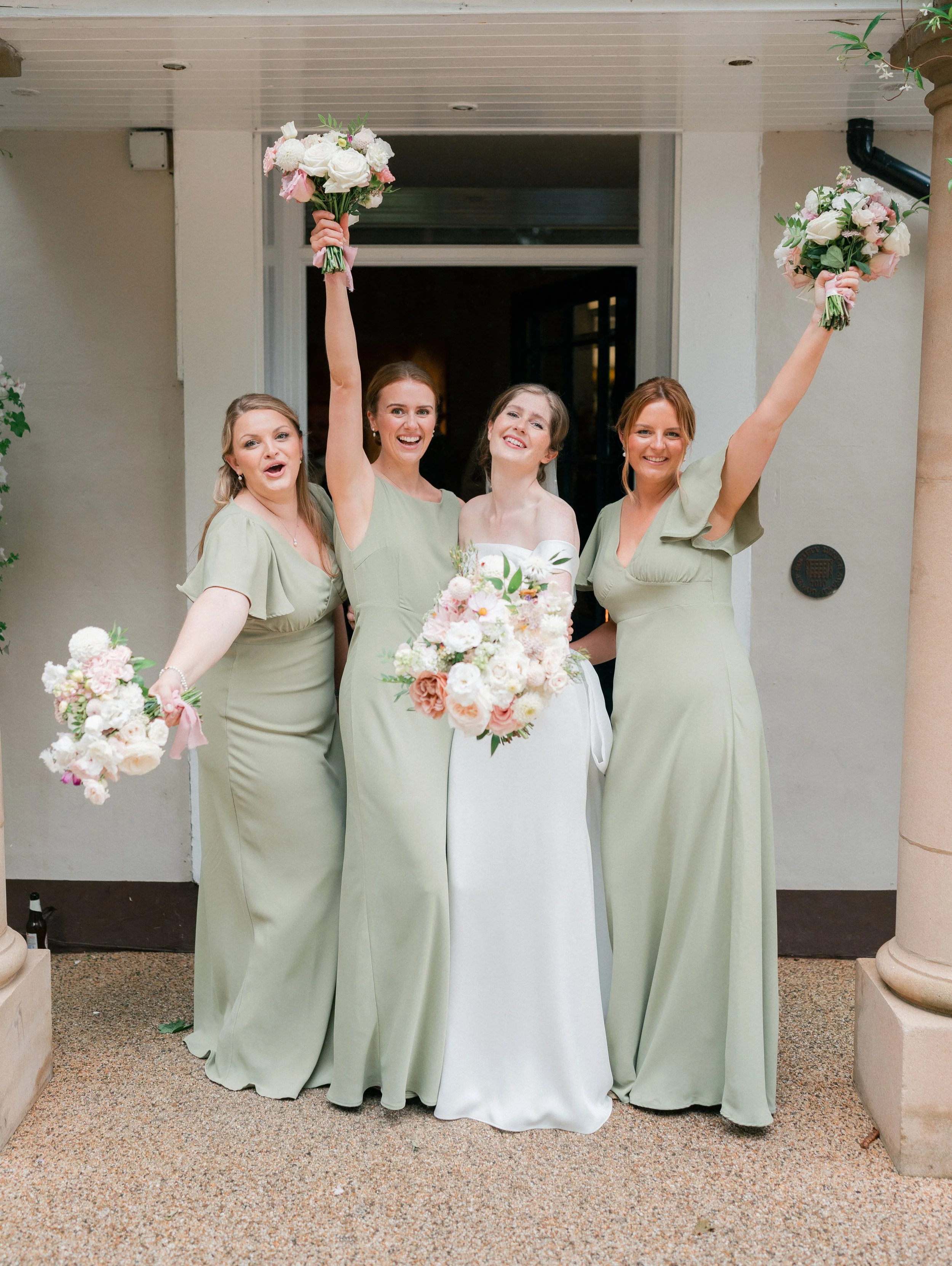 Four women, including a bride and bridesmaids, celebrating with bouquets of pink and white flowers outdoors.