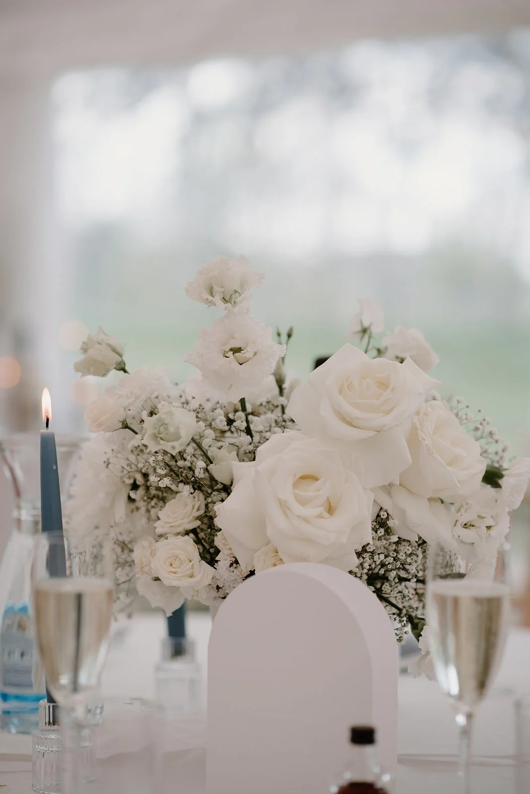 small bowl of flowers on a wedding reception table, white roses and smaller white flowers with a little gypsophillia, glass of champagne in the foreground and blue tapered candles in the background. 