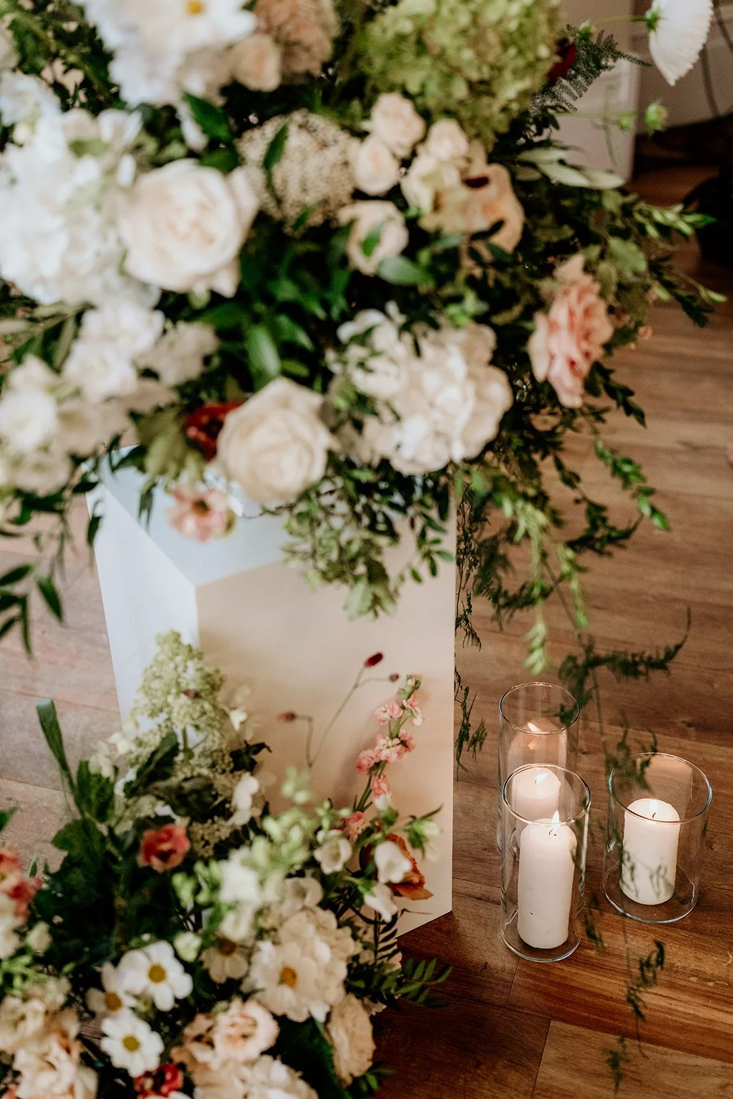 Close-up of a floral arrangement with white and light pink flowers, greenery, and candles on a wooden floor.