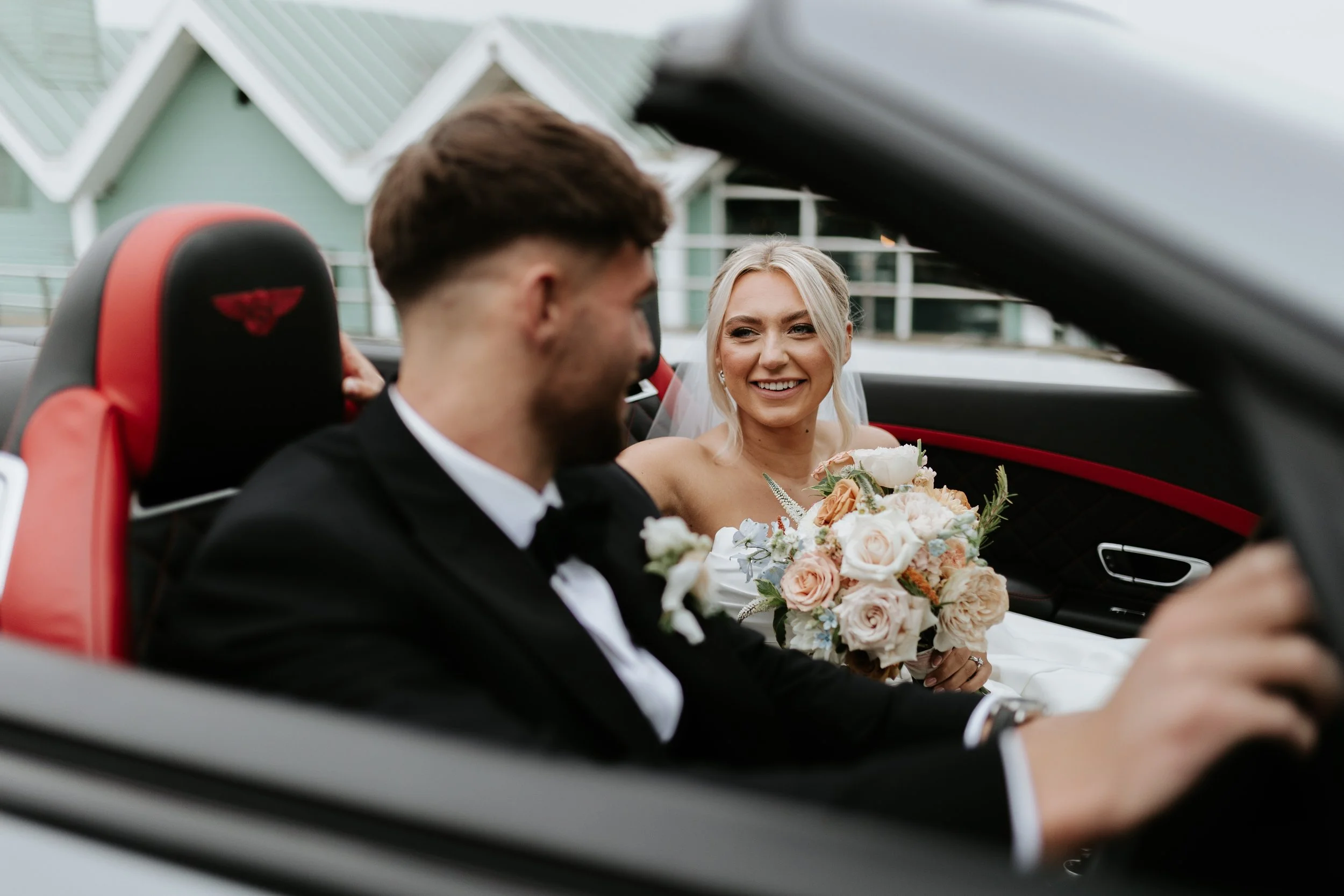 A bride and groom sitting in a convertible car, with the bride smiling at the groom. The bride is holding a bouquet of flowers, and the groom is wearing a tuxedo. The interior of the car is black and red, and the background shows modern houses.