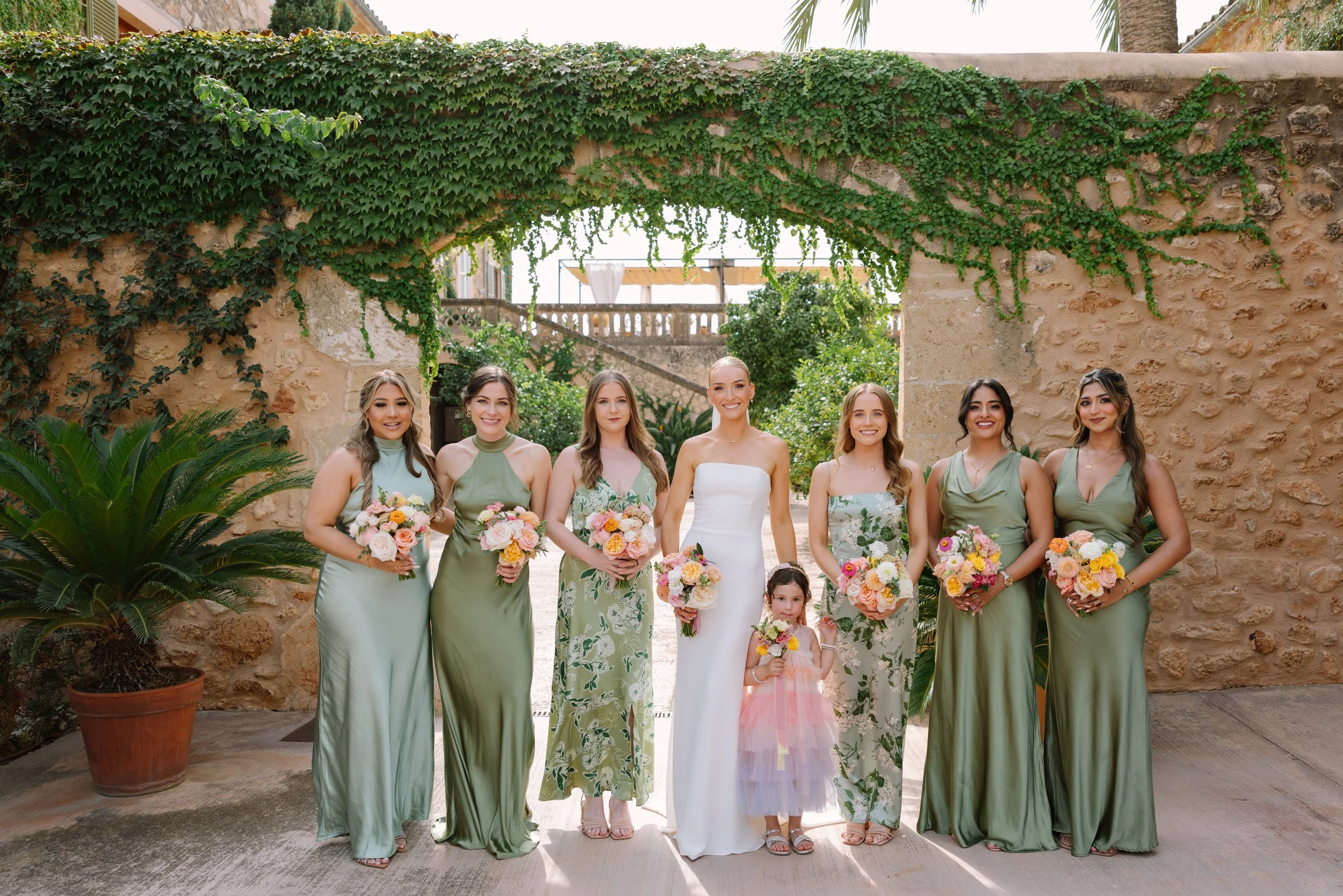A group of eight women and one girl standing outdoors in front of a stone wall with greenery, dressed in formal attire, holding bouquets of flowers, smiling for a wedding or special occasion photo.