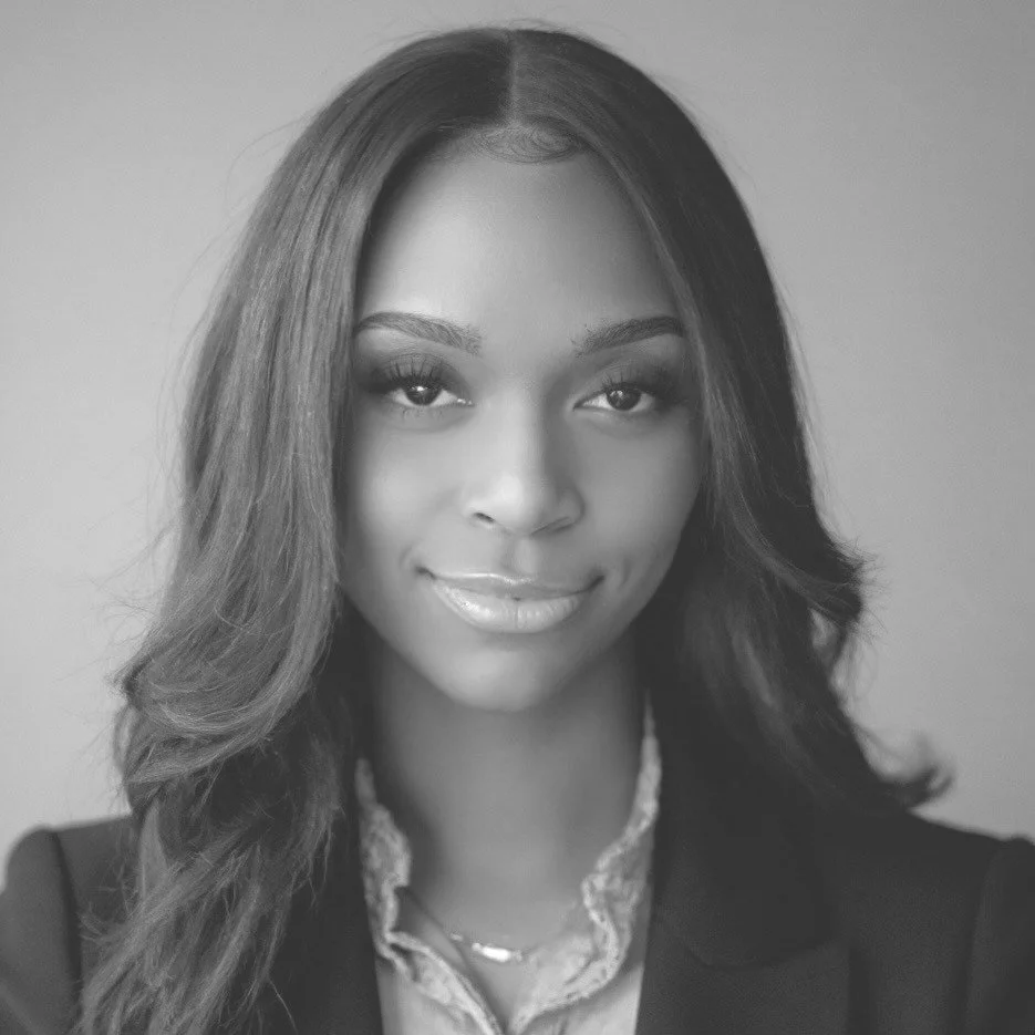 Black and white portrait of a young woman with long, wavy hair, smiling softly, wearing a blazer and a necklace.