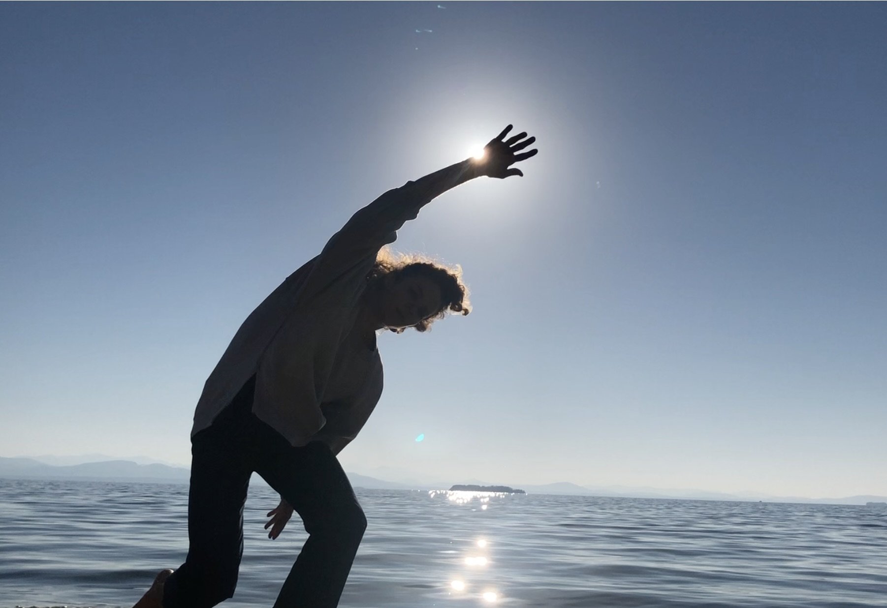 Jocelyn Tobias, founder of Seachange Conversations, silhouetted in front of a large calm body of water, reaching upward with her hand just in front of the sun, reflecting her dance background and the light of lived experience.