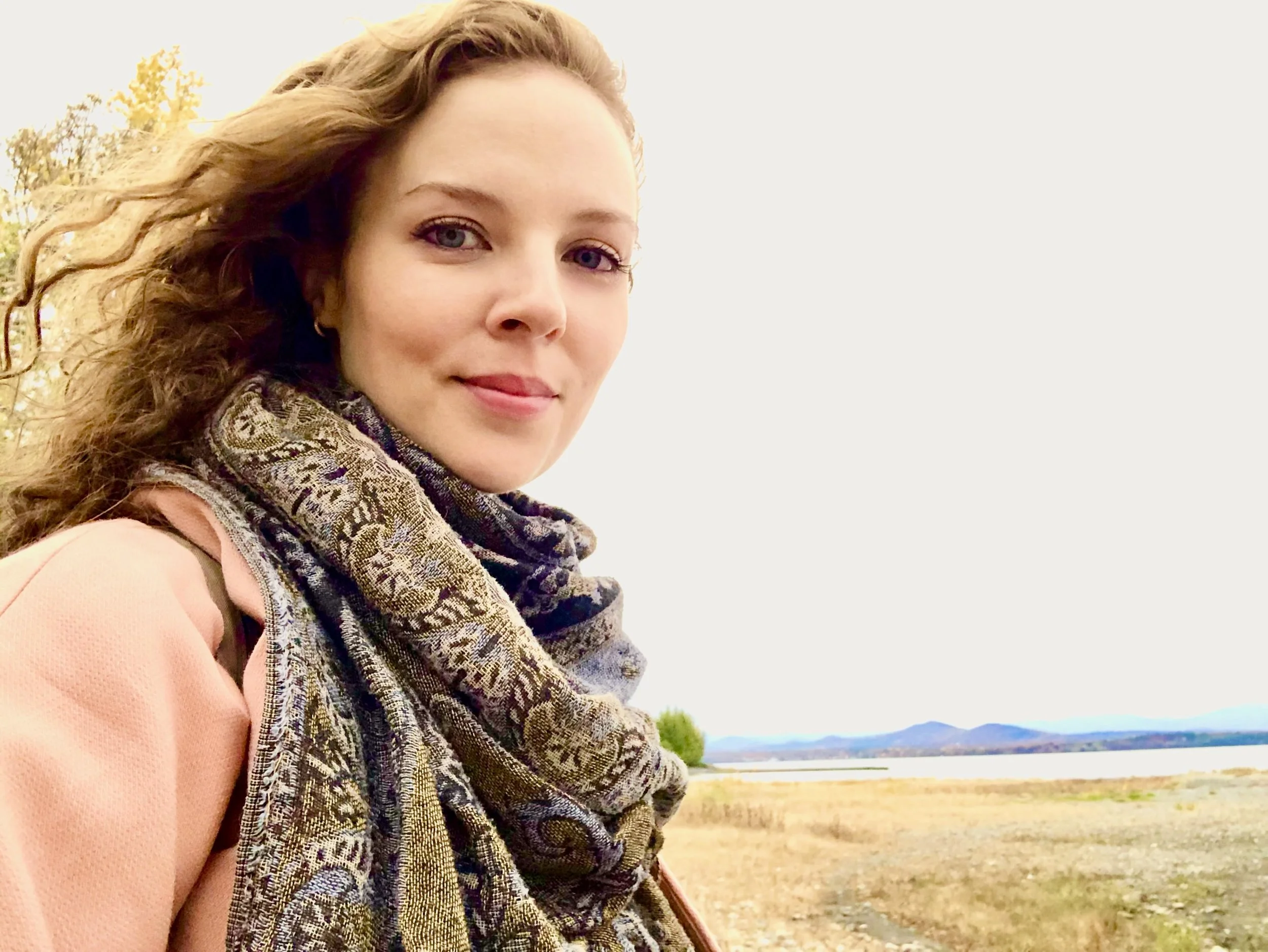 A woman with long curly hair standing outdoors near a body of water, wearing a patterned scarf and a peach-colored jacket, with a background of water, trees, and distant mountains.
