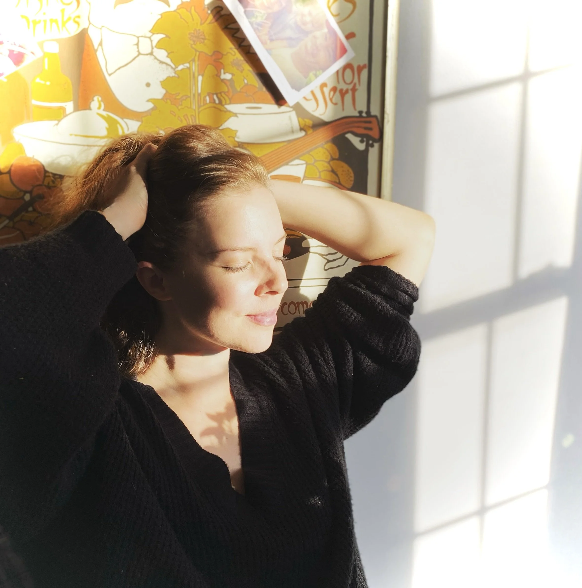 A woman relaxed with her eyes closed, basking in sunlight, against a colorful, illustrated wall with a window casting light and shadow.