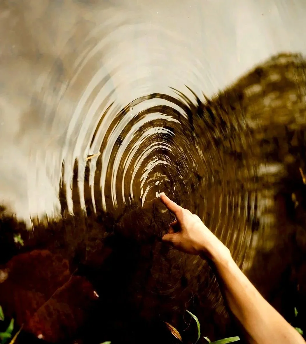 A person's pointer finger connecting with the  surface of a small body of water, creating a ripple effect. Image in turned sideways so the water can also read as a mirror.