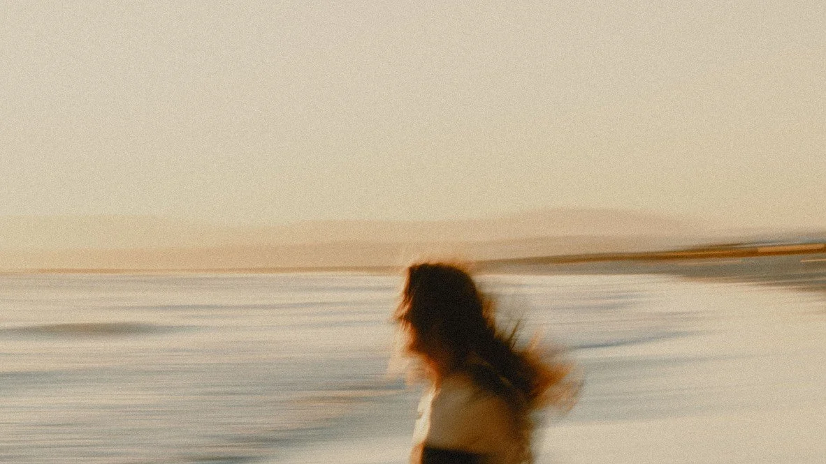 Blurred image of a person with long hair standing near a body of water at sunset, contemplative and still, representing the quiet moments of realization when something is ready to change.