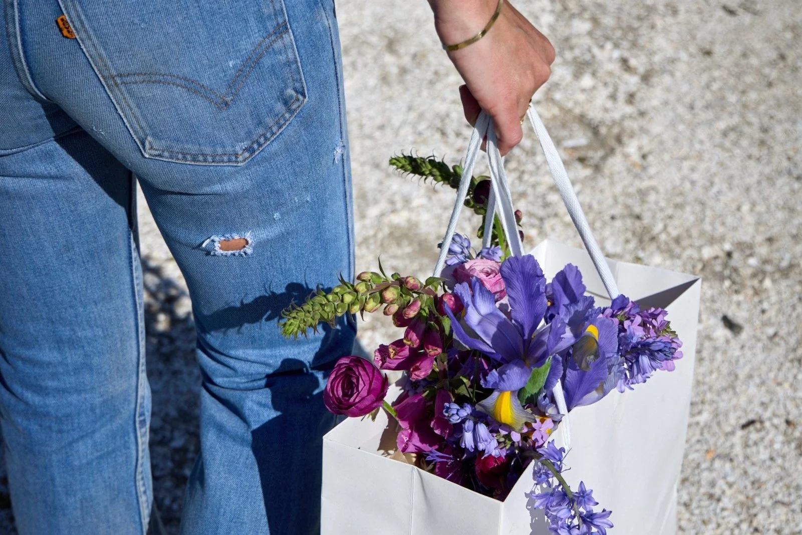 Person in blue jeans holding a paper bag with purple, pink, and yellow flowers inside, standing on a gravel surface.