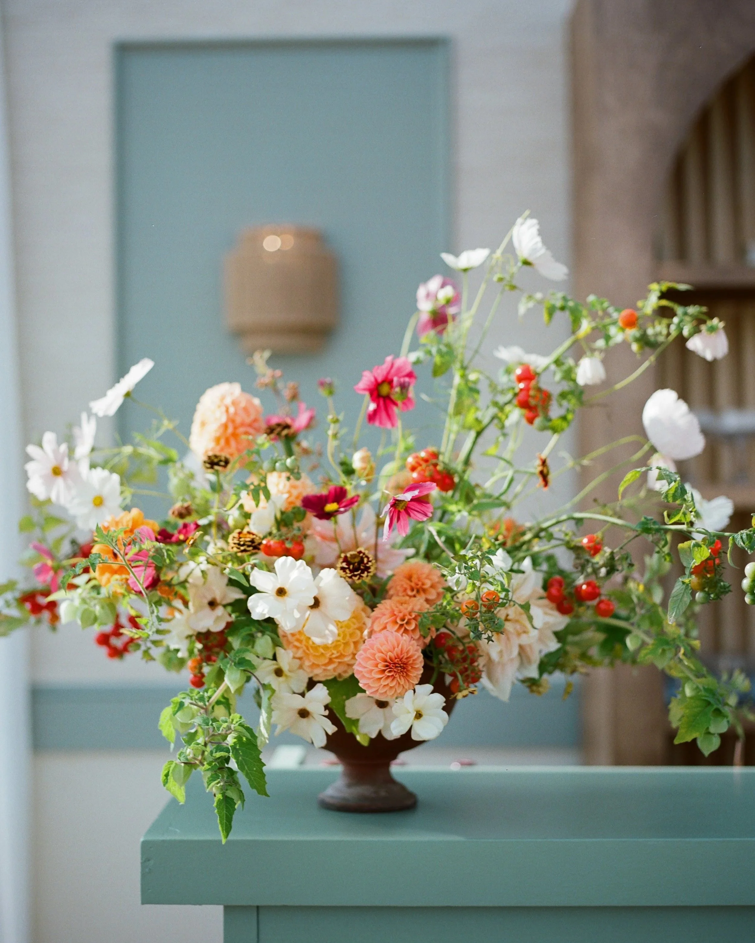 A large bouquet of colorful flowers in a brown vase on a teal table in a room with pale walls and a blue doorway.