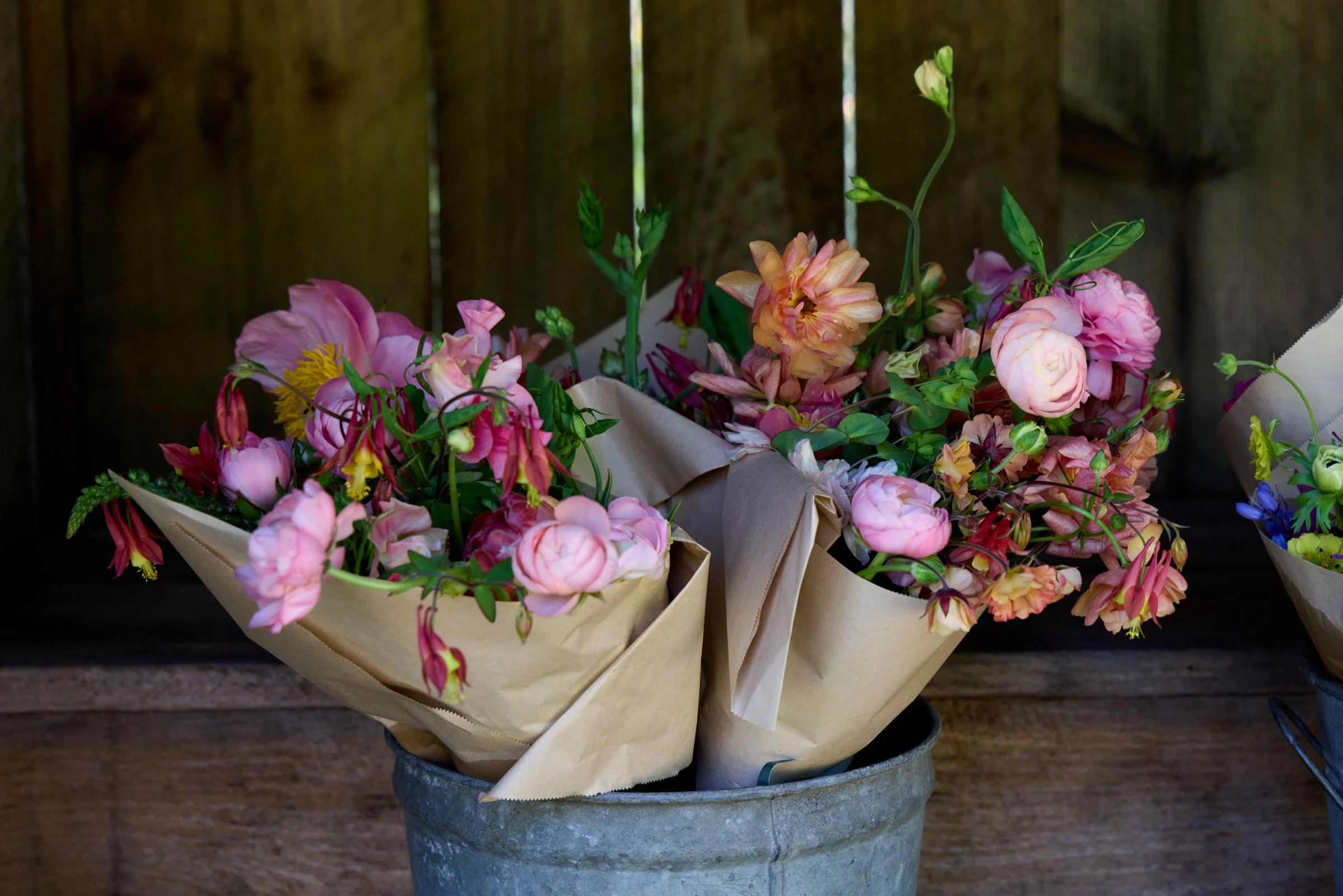 Pink and orange flowers in paper wrapping in a metal bucket with a wooden background.
