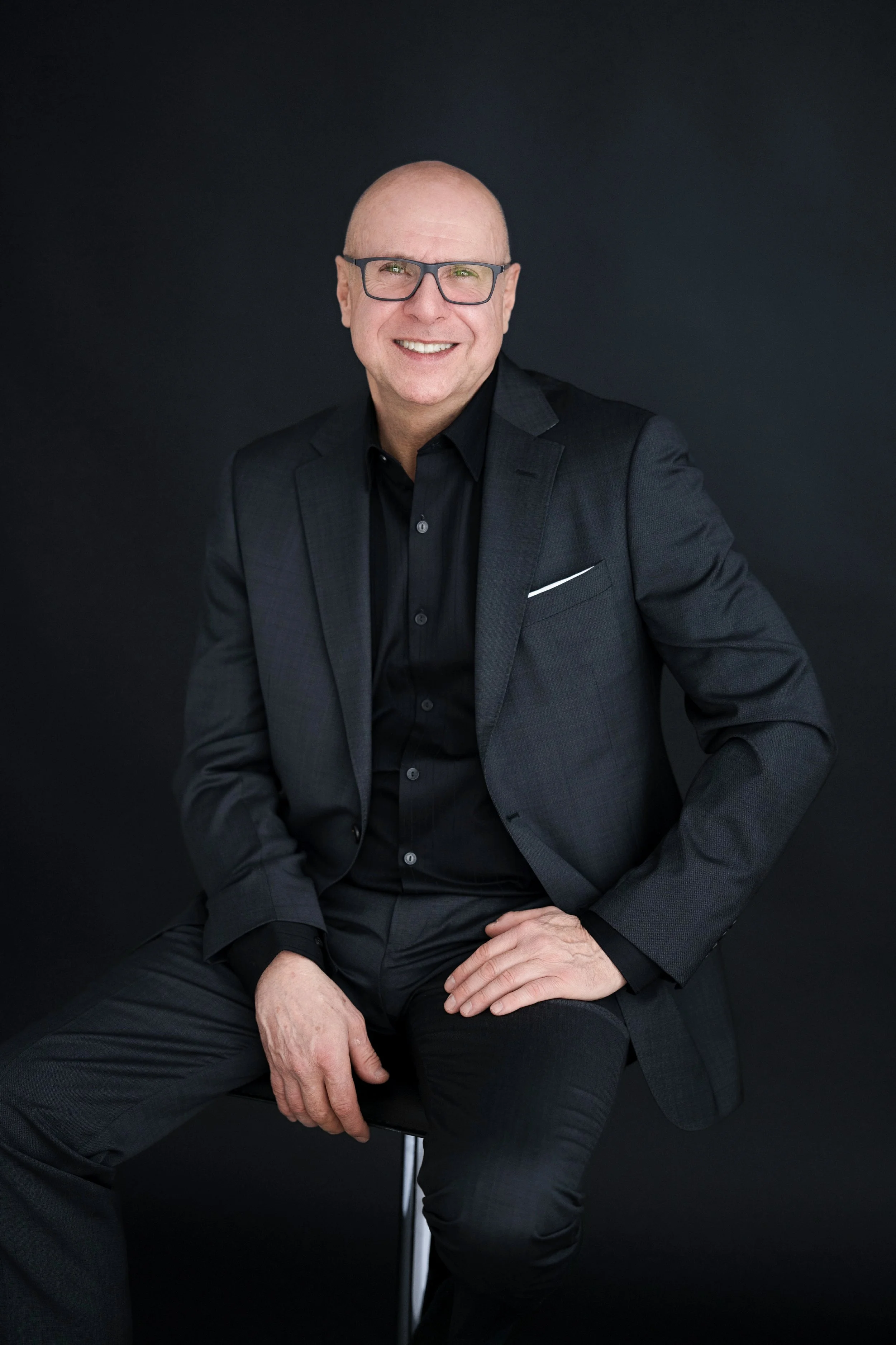 Homme en costume noir assis sur une chaise, souriant, portant des lunettes, fond noir.