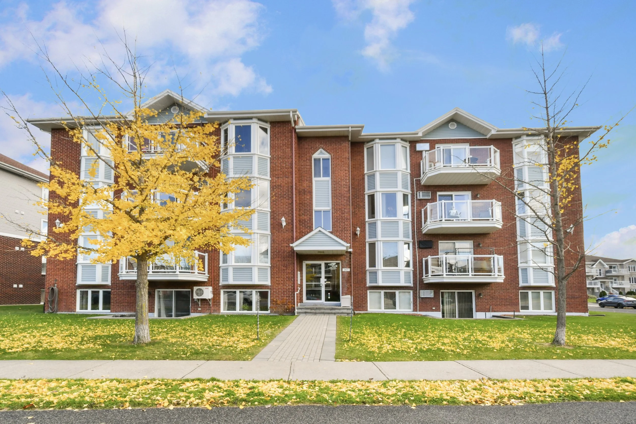 Un immeuble résidentiel en briques rouges avec plusieurs balcons et grandes fenêtres, entouré d'arbres dont certains ont des feuilles jaunes en automne, sur un fond de ciel bleu avec quelques nuages.