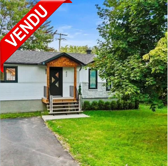 Maison moderne avec entrée en bois et escalier, entourée d'arbres et de verdure, avec une étiquette 'VENDU' en haut à gauche.