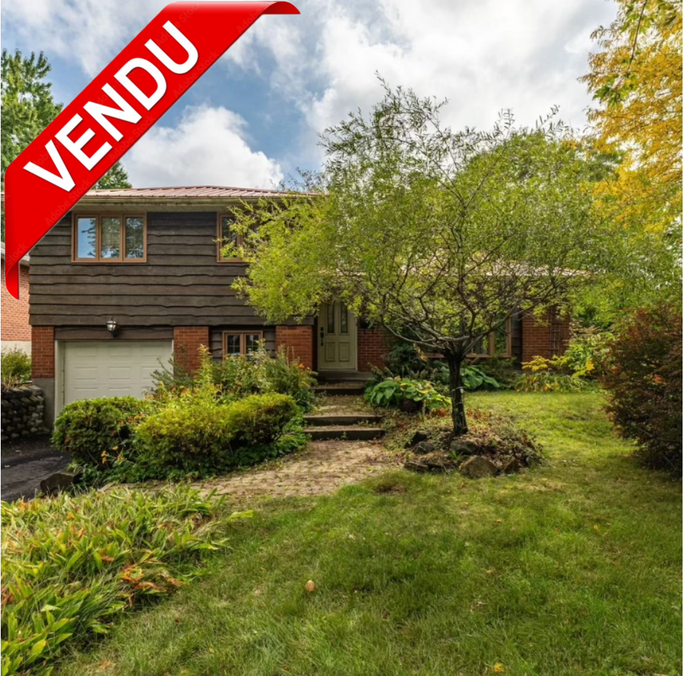 Maison avec garage, entourée d'un jardin verde avec un arbre, des arbustes et un chemin de pierre, ciel partiellement nuageux, image avec un bandeau rouge avec la mention 'VENDU' en français.