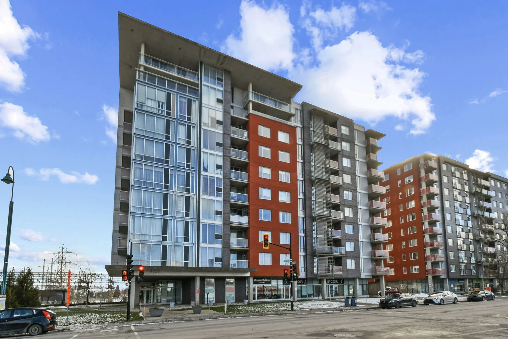 Immeuble résidentiel moderne avec plusieurs étages, façades en verre et couleurs vives, situé le long d'une rue avec plusieurs voitures stationnées, un feu de circulation rouge et un ciel ensoleillé avec quelques nuages.