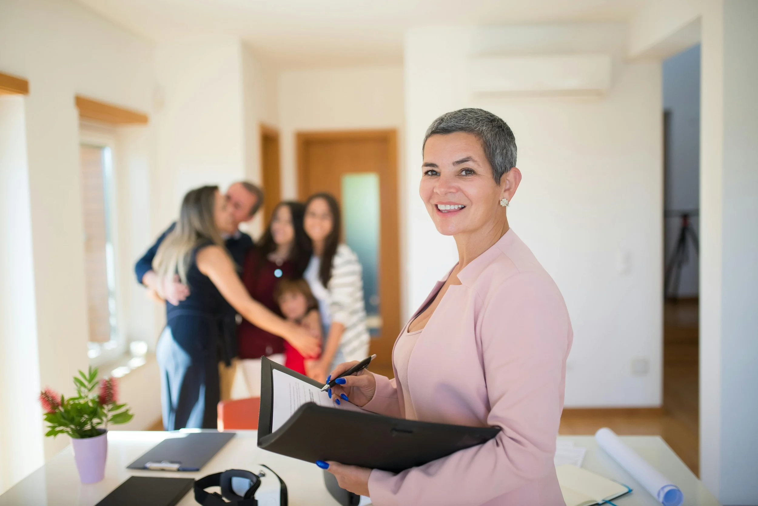 Une femme souriante tenant un carnet, dans un bureau, avec un groupe de personnes en train de se faire un câlin en arrière-plan.