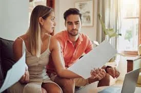 Un couple assis sur un canapé, examinant des papiers dans un salon bien éclairé.