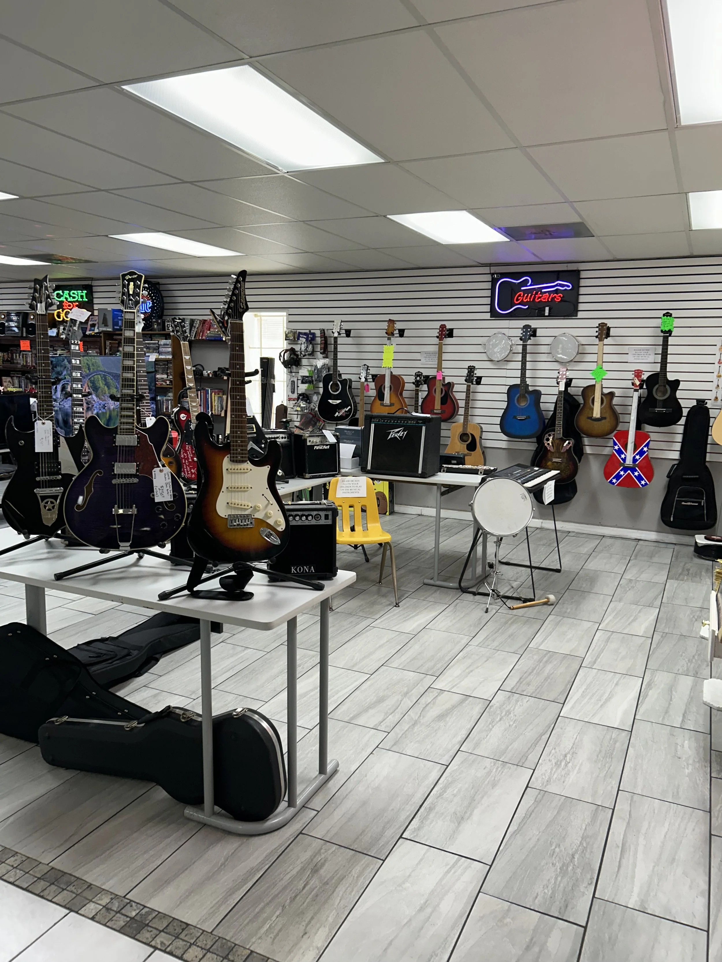 A music store interior displaying guitars hanging on the wall, with some electric guitars on a table in the foreground, alongside guitar cases on the floor. There are also musical instruments like drums, amplifiers, and signage neon signs for guitars and cash. The store has white tiled flooring and a slatwall display.