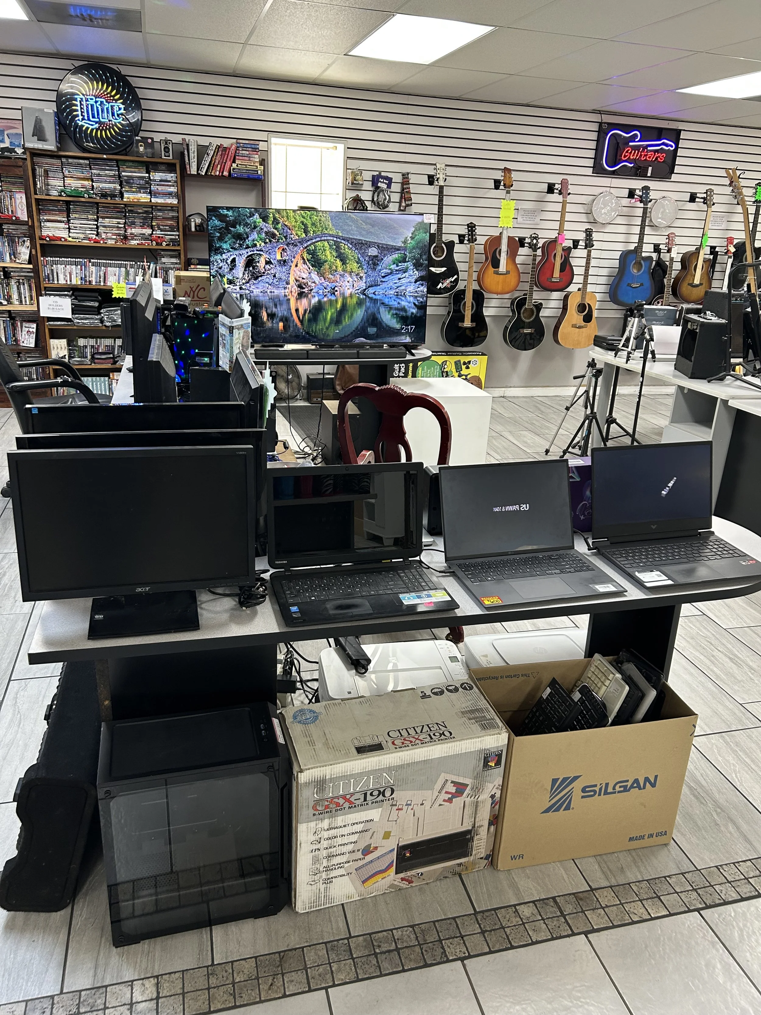 An indoor store with guitars displayed on the wall, a large TV showing a scenic image, multiple computer monitors and laptops on tables, with boxes of printers and accessories in the foreground, and books and DVDs on shelves on the left wall.