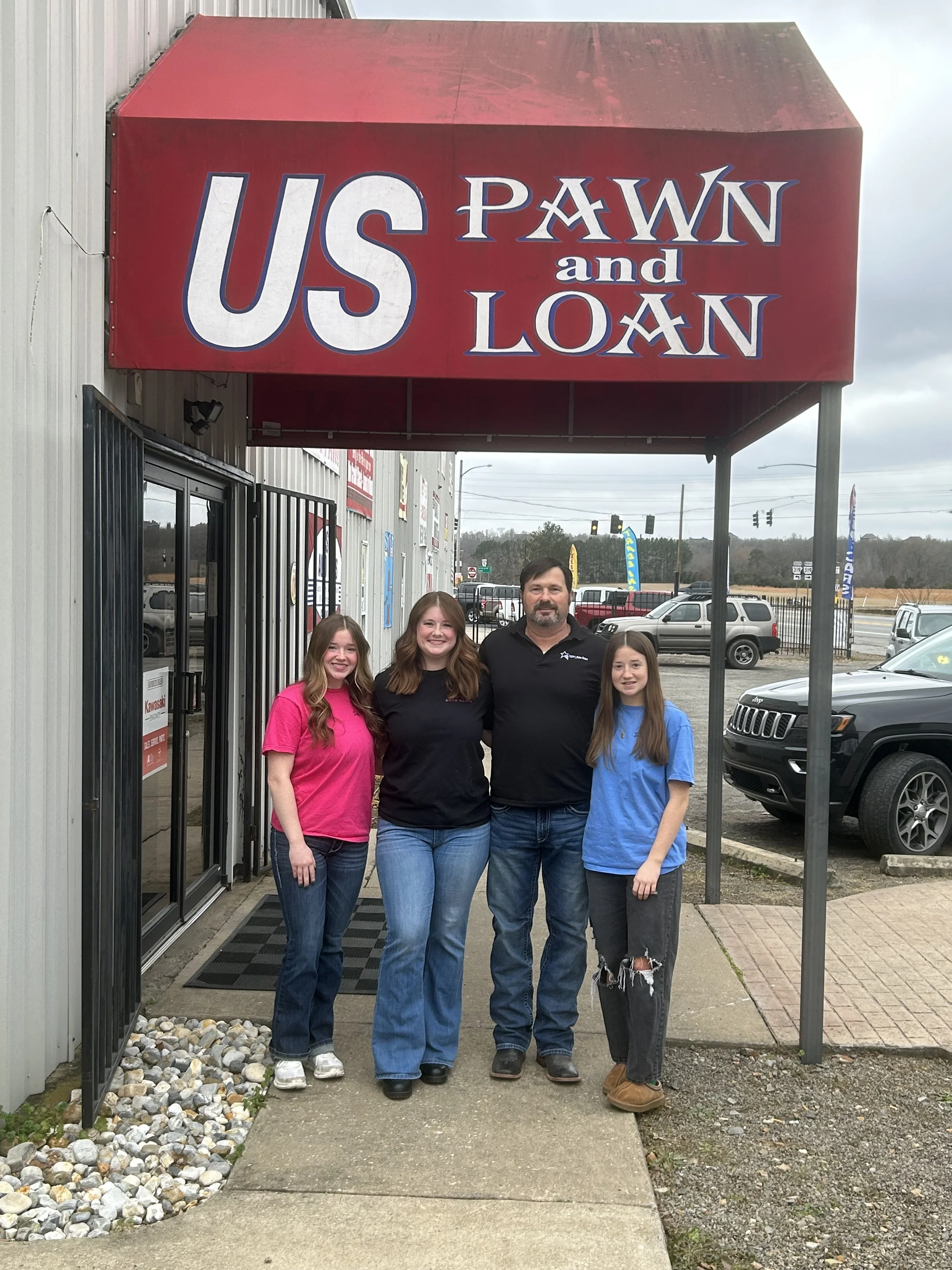 Group of four people standing outside the U.S. Pawn and Loan store under a red awning, with a parking lot and cars in the background.