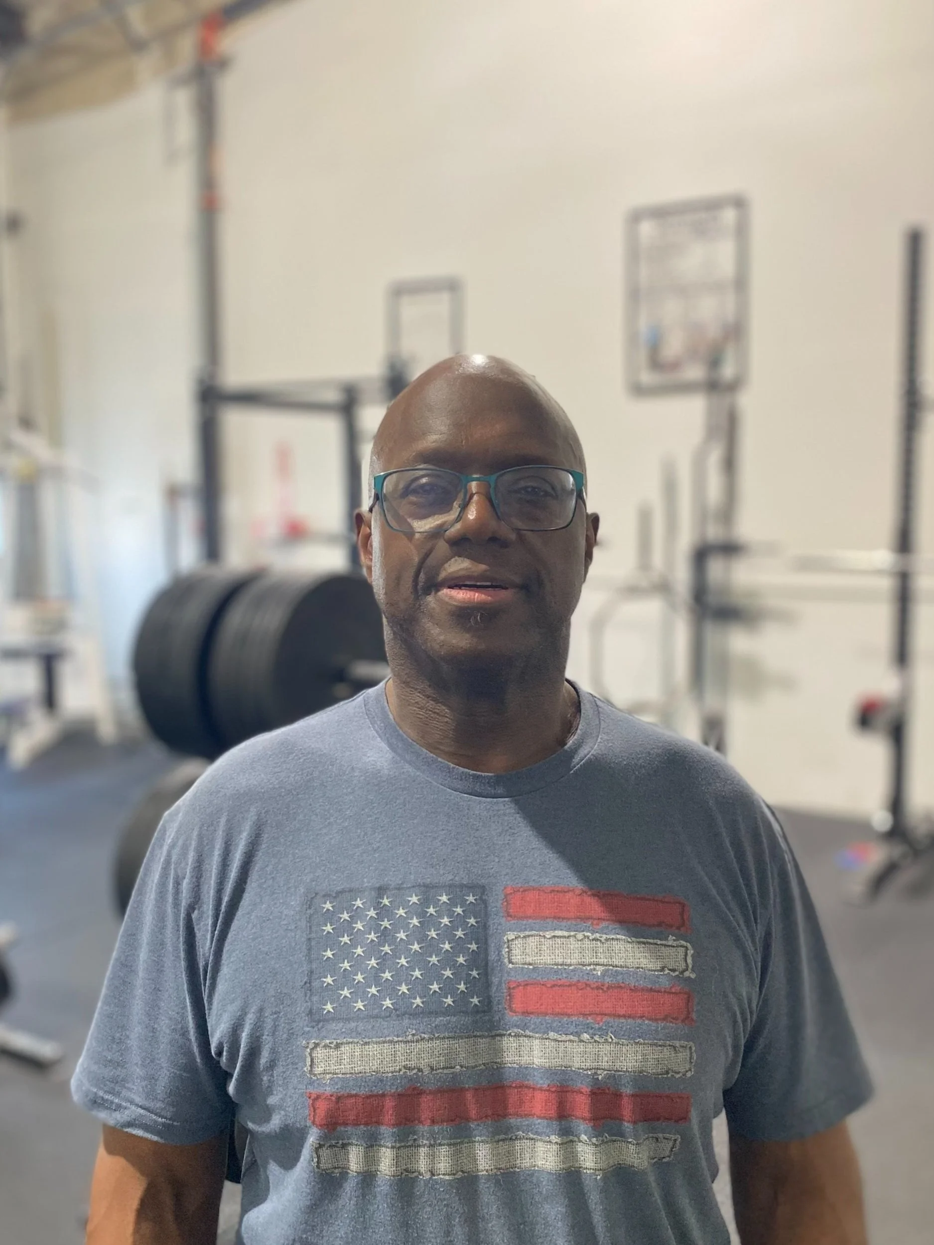 A man with glasses standing in a gym, wearing a t-shirt with an American flag design.