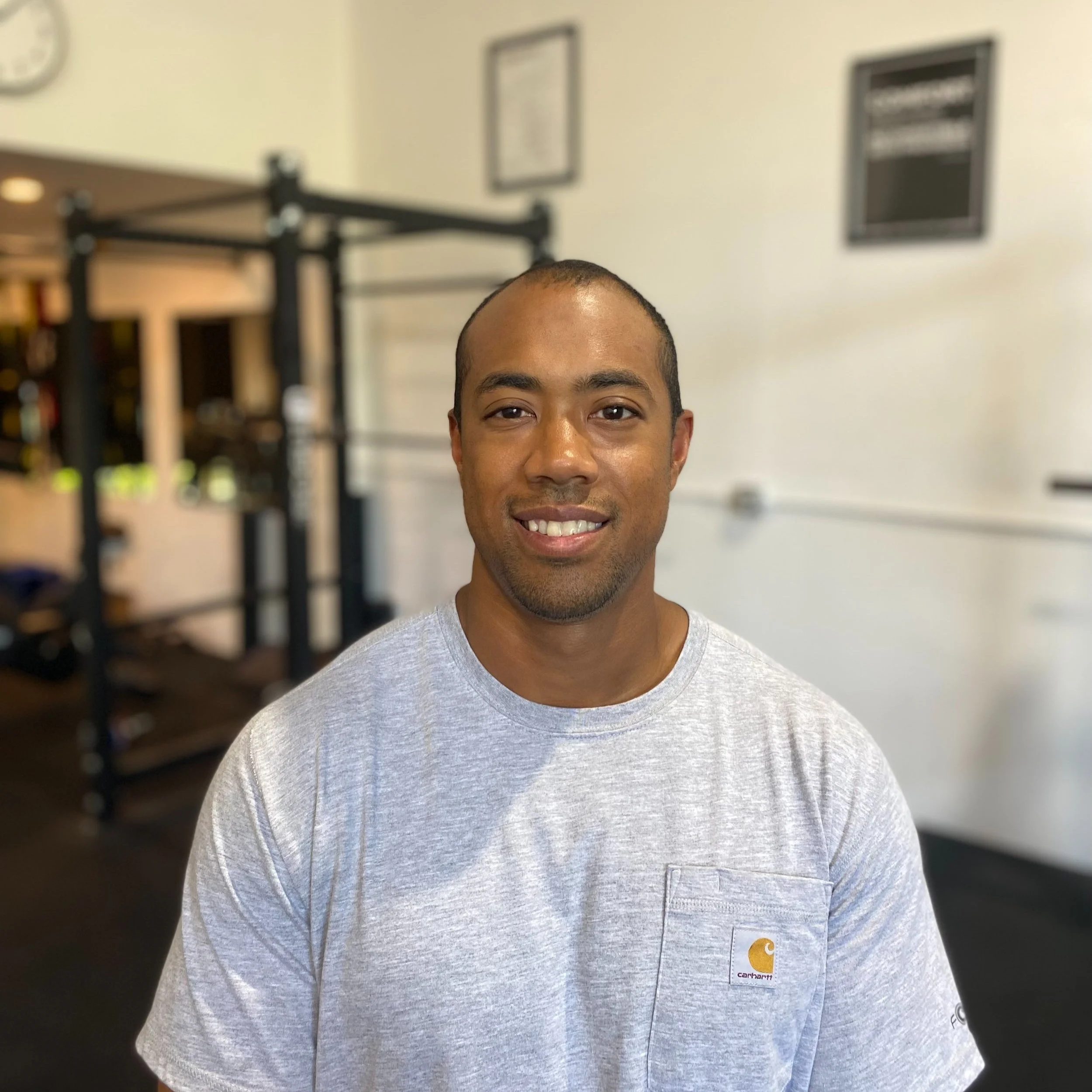 A smiling man with medium skin tone and short hair, wearing a light gray Carhartt t-shirt, standing inside a gym.