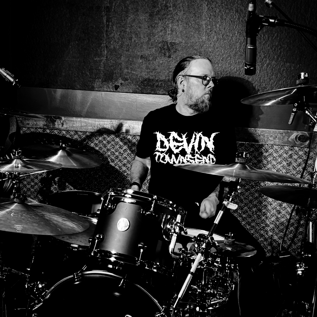 Black and white photo of a man with glasses and a beard playing the drums in a recording studio, wearing a T-shirt with a graphic logo.
