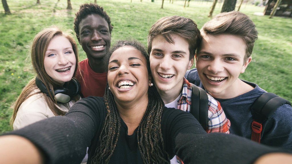 Group of five teenagers taking a selfie together outdoors in a park.