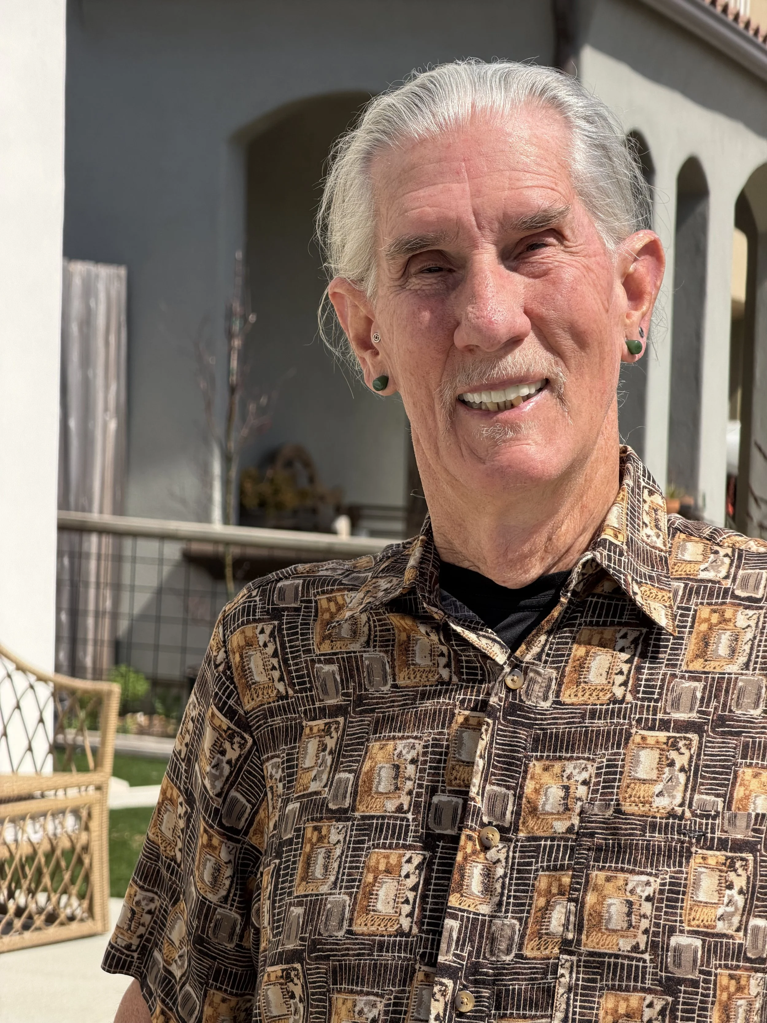 An older man with gray hair, earrings, and a patterned shirt standing outside in a sunny yard.