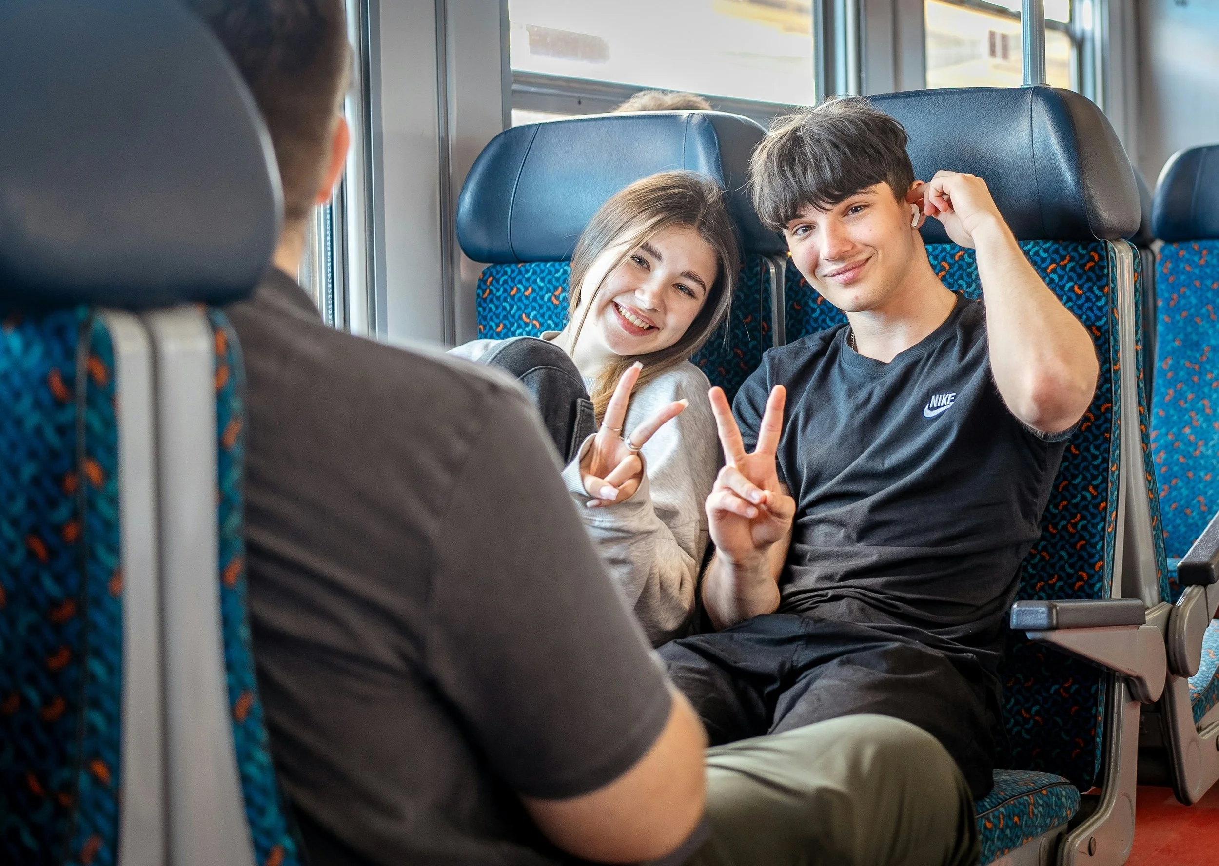 Two teenagers, a girl and a boy, sitting in a vehicle and smiling at the camera, making peace signs, with a person in the foreground.