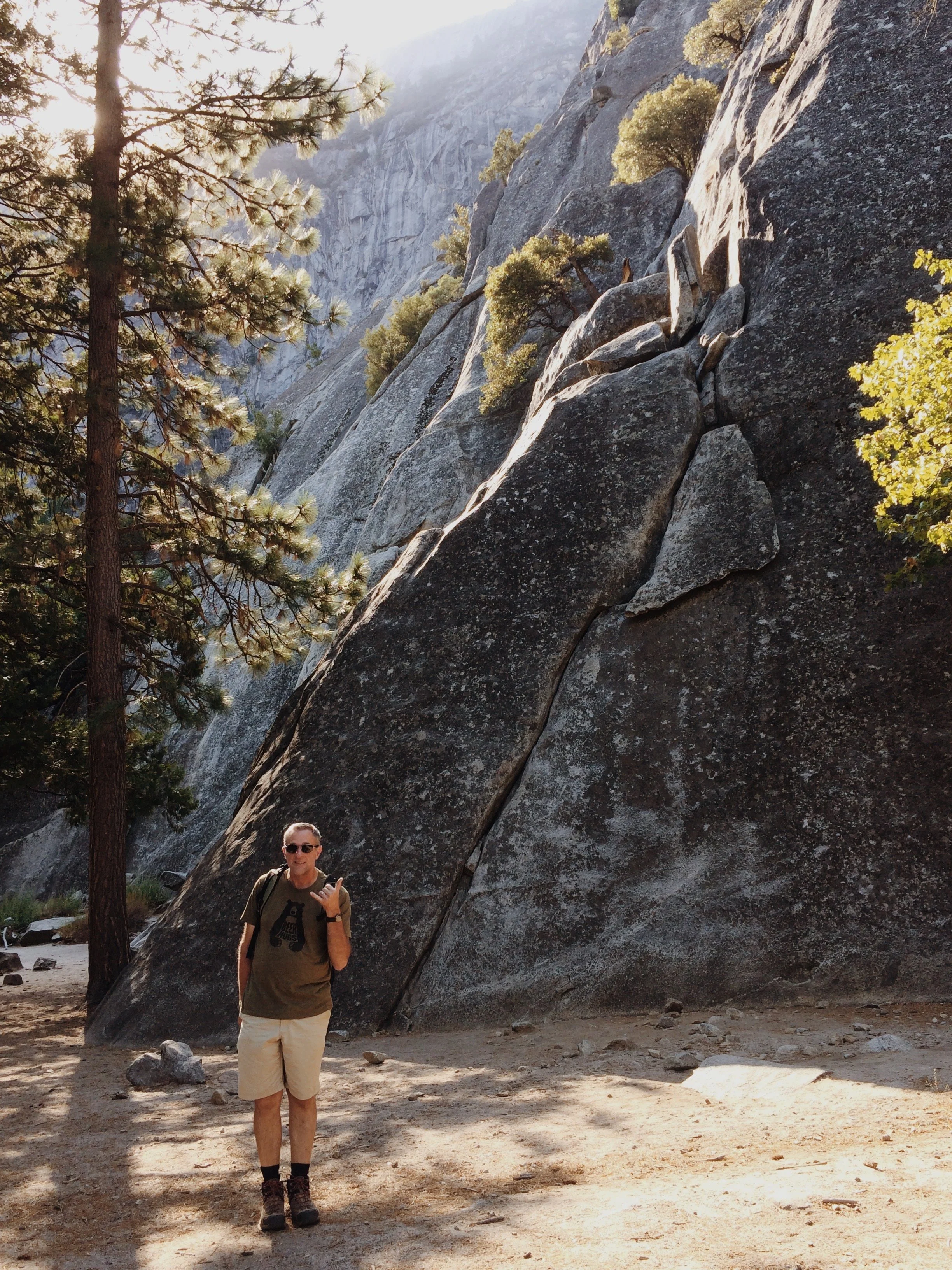 A man standing in front of a large granite rock formation, holding a phone and giving a thumbs-up, with trees and mountains in the background.