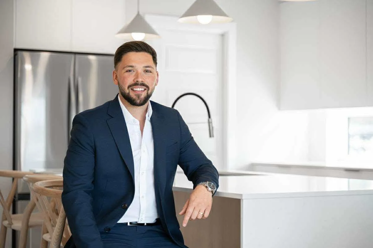 Un homme en costume bleu assis dans une cuisine moderne avec un fond blanc et un évier noir.