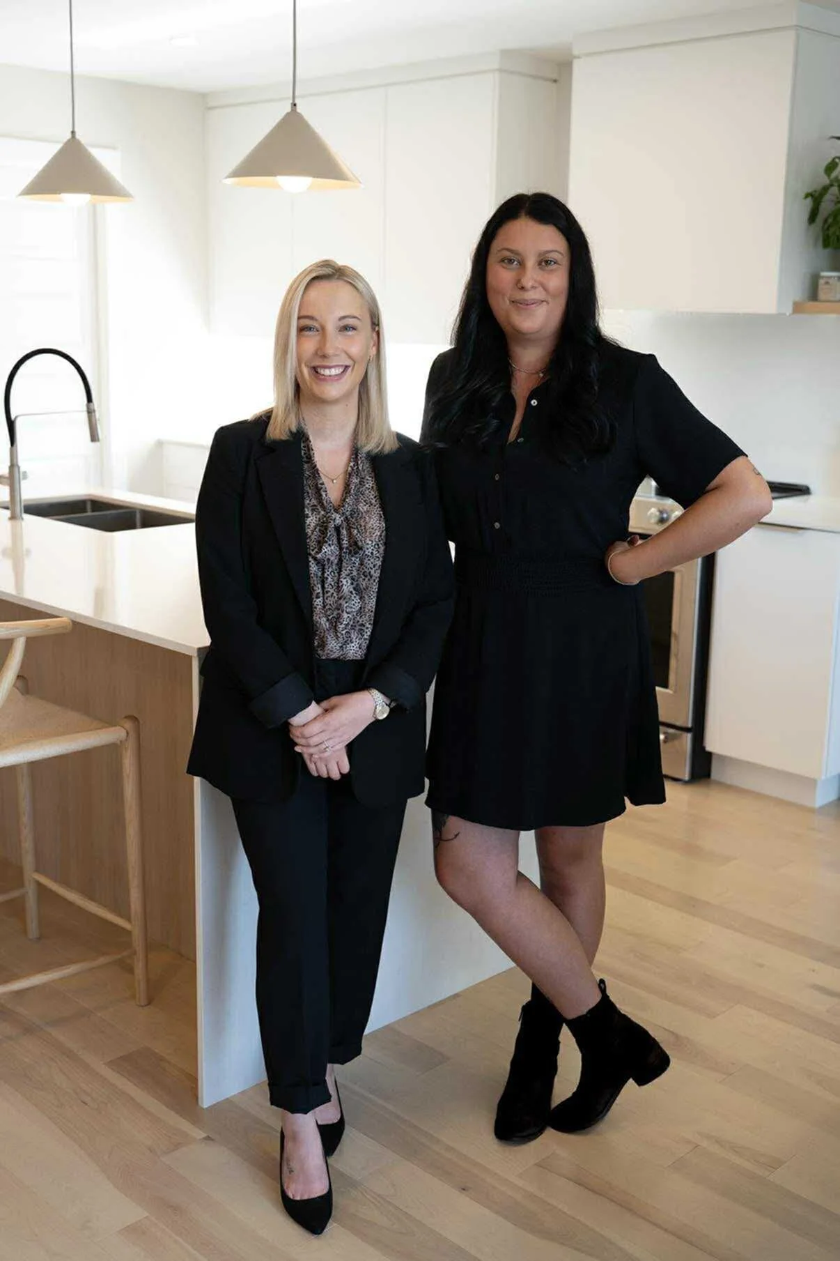 Deux femmes posent dans une cuisine moderne, l'une habillée en tailleur noir avec un chemisier à motifs, l'autre en robe noire avec des bottines noires.
