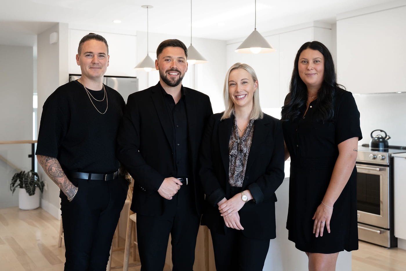 Quatre personnes professionnelles, deux hommes et deux femmes, habillées formellement, posant dans une cuisine moderne avec des appareils en acier inoxydable et un éclairage suspendu.