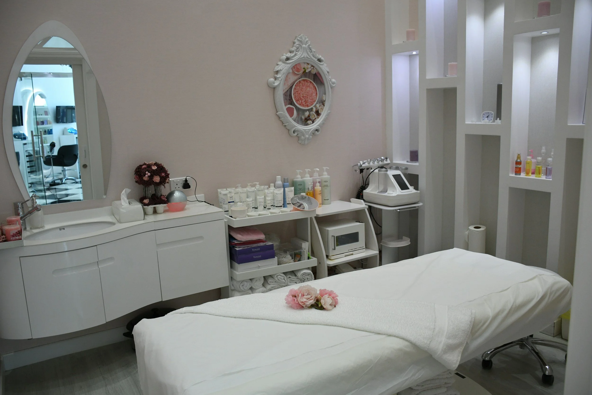 A massage or treatment room with a bed covered in white sheets and a towel with pink flowers on top, a white vanity with a mirror, shelves with skincare products, and a clinical device, all in a clean, brightly lit space with pastel decor.