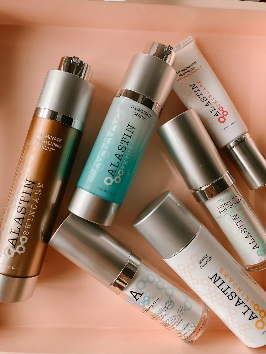 Various skincare products from the brand Alastin, including serums, a cleanser, and a skin revitalizer, arranged on a light pink surface.