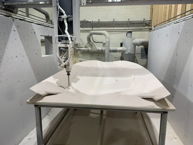 Industrial 3D printing machine creating a large, white, complex sculpture resembling a futuristic vehicle or craft on a metal table in a manufacturing facility.
